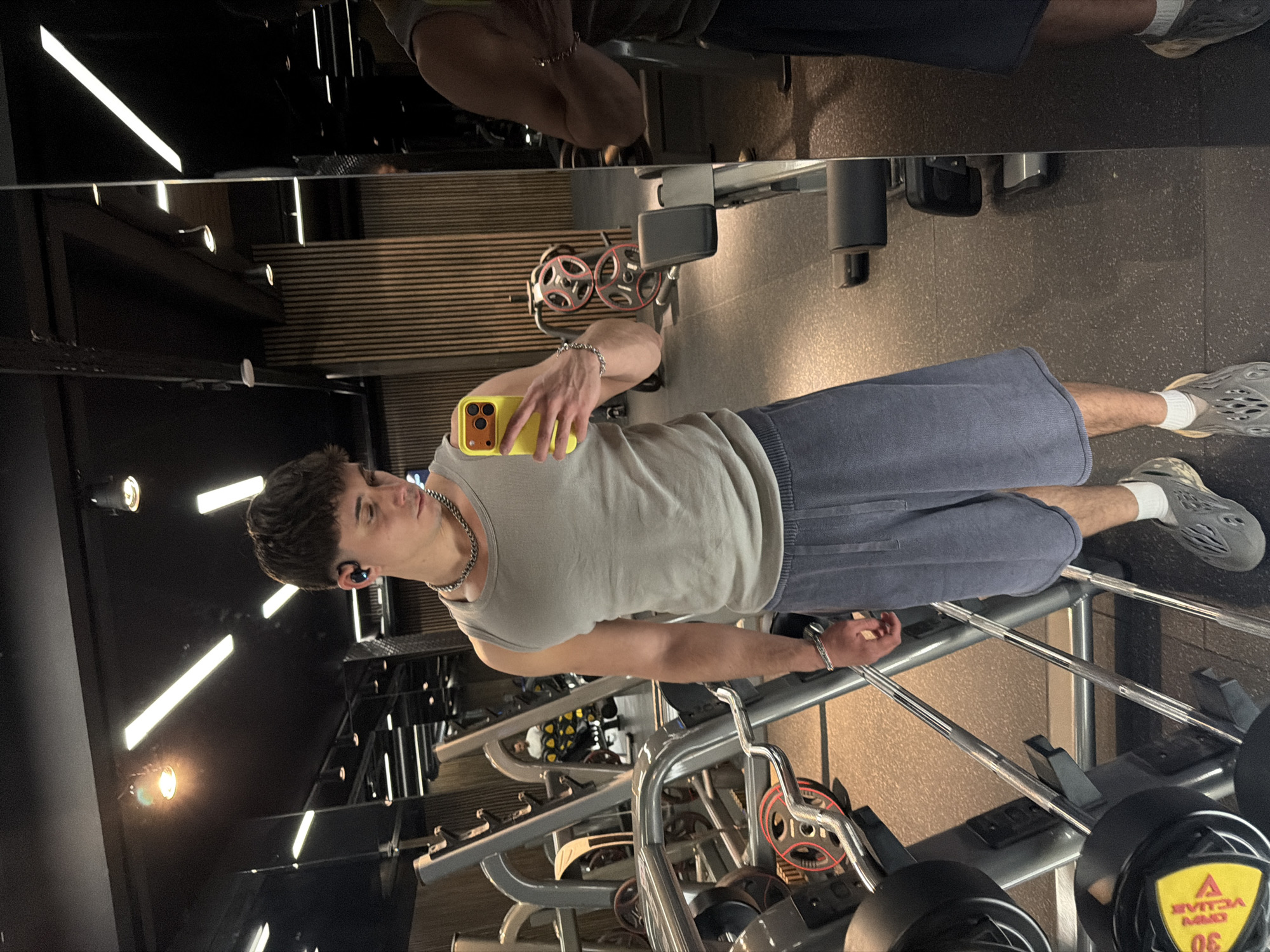 A young adult male is captured in a full-body mirror selfie taken inside a gym. He stands facing forward, holding a yellow-cased smartphone in his left hand to photograph his reflection. The male has short brown hair and wears a light grey tank top, dark blue athletic shorts, white socks, and grey perforated slip-on shoes. Accessories include black wireless earbuds in both ears, a silver chain necklace, and a silver chain bracelet on his left wrist. The gym environment features dark grey rubber flooring, black ceilings with linear and spot lighting, and walls composed of large mirrors and vertical wooden slats. Various pieces of exercise equipment, including weight racks, barbells, and plate weights (one marked '30 KG'), are visible throughout the space. A blurred reflection of another individual in a white t-shirt is faintly visible deeper within the gym, reflected in the mirror. No specific details within the image allow for the determination of its location within Sofia, Bulgaria.