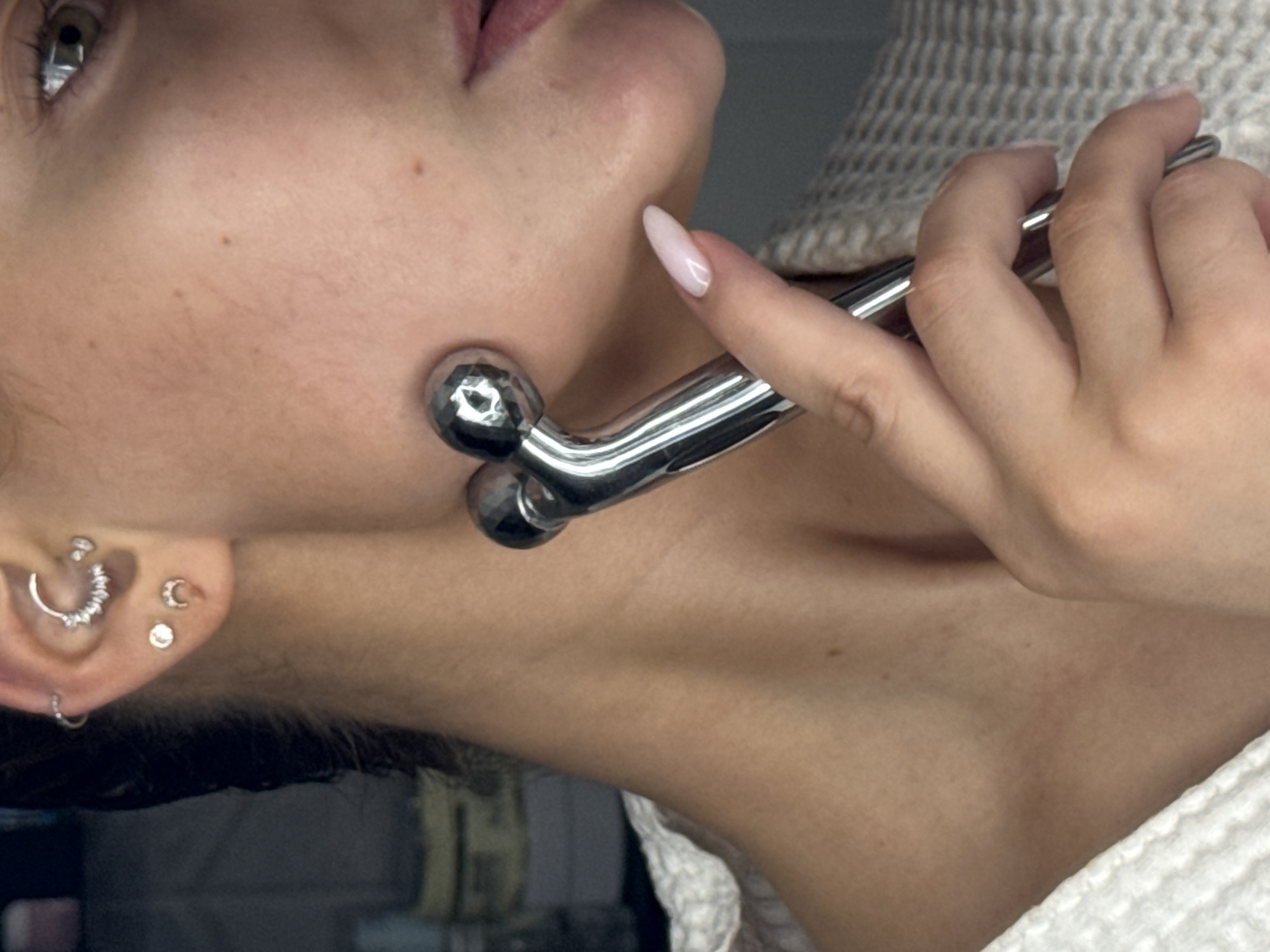 The image depicts a person using a metallic facial roller on their cheek. The person's left ear is visible, adorned with multiple piercings. The person's hand is holding the facial roller, and the thumb has a light pink manicured nail. A light-colored, textured garment is draped around the person's shoulder. The location is not discernible from the image, and there is no indication that the image was taken in Chaikovsky, Russia.