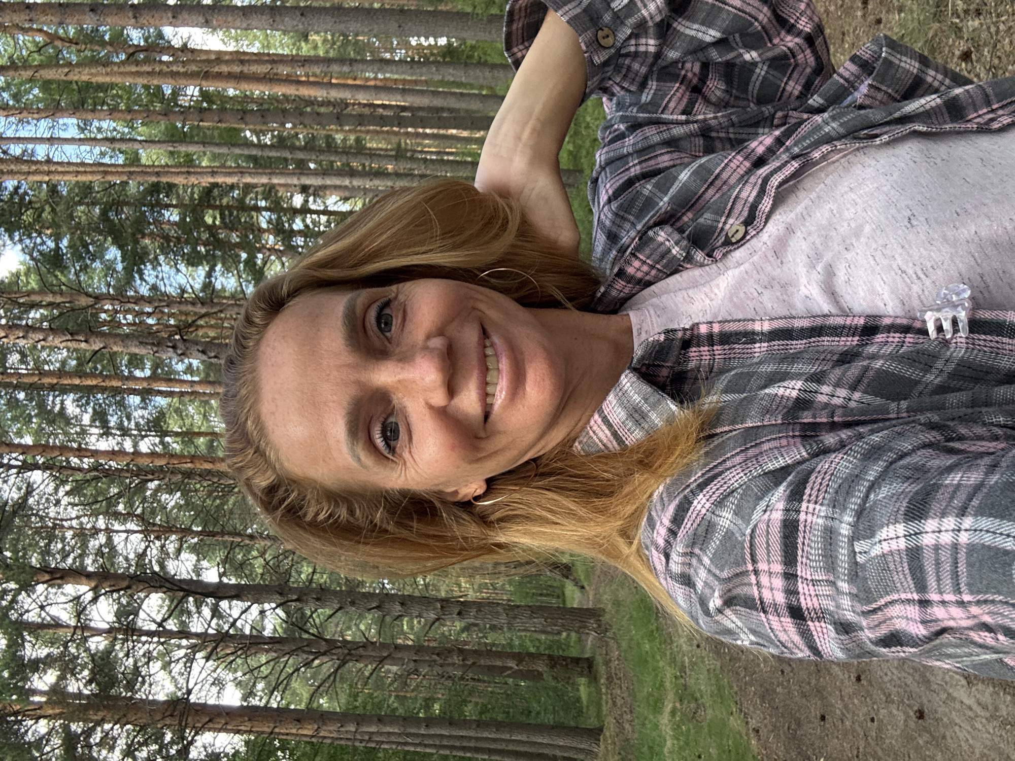A woman with auburn hair and freckles smiles at the camera, her right arm resting behind her head. She is wearing a pink and grey plaid shirt over a pink t-shirt, and a small, clear plastic hair clip is attached to the t-shirt. The background consists of a dense forest of tall pine trees, with a dirt path visible in the foreground. The scene is set in the city of Portovoe, Russia.