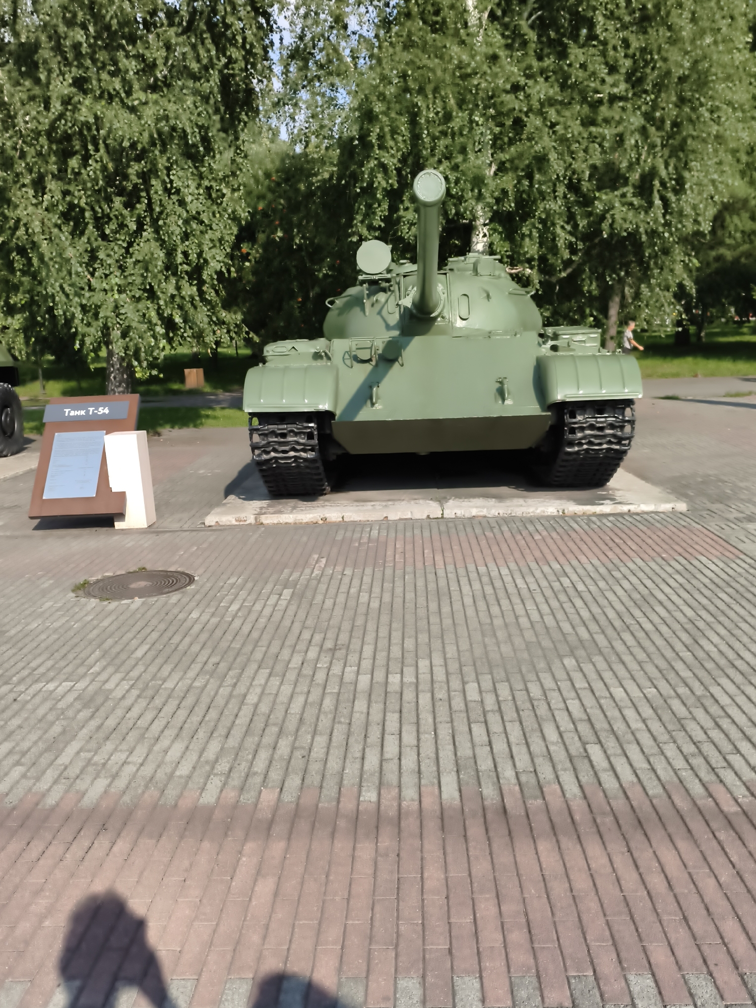 The image shows a green tank, labeled "Танк Т-54" (Tank T-54), displayed on a paved area in a park-like setting. The tank is the central object, facing forward with its turret and gun visible. It sits on a concrete base, with its tracks resting on the paving stones. To the left of the tank, there is a brown display board with a white section, providing information about the tank.
The setting appears to be an outdoor park or memorial site, with lush green trees in the background and a clear blue sky. The ground is paved with interlocking stones, a mix of gray and reddish-brown. The lighting suggests it is daytime, likely a sunny day, as indicated by the shadows cast on the ground, including what appears to be the shadow of the photographer and possibly another person.
In the background, a few people are visible, including a child running. The overall atmosphere seems peaceful and perhaps commemorative, given the display of the historical tank. There are no strong emotional cues conveyed, but the scene suggests a public space for historical display and recreation. The visible text confirms the object is a T-54 tank, and the location context indicates Chelyabinsk, Russia.