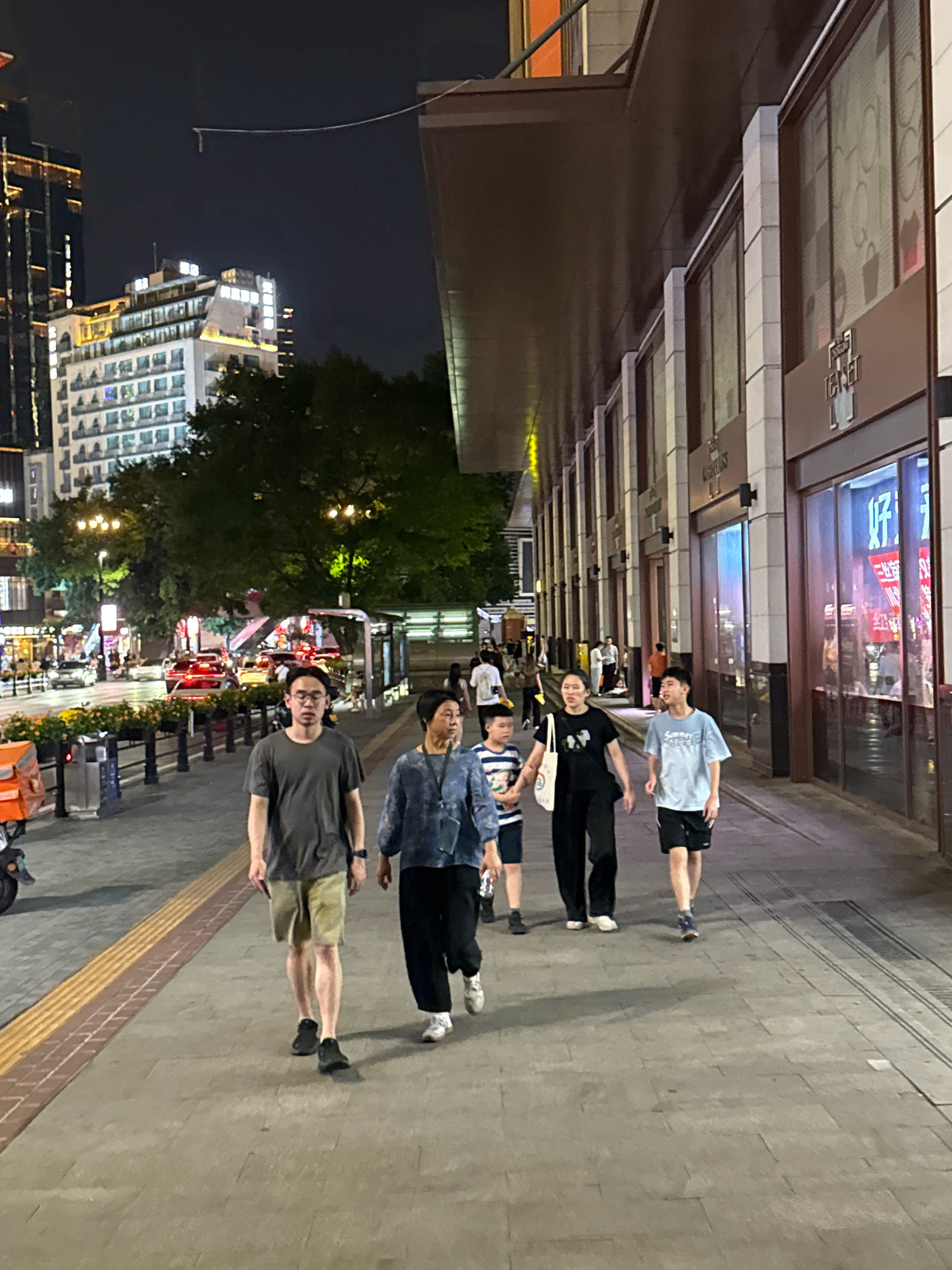 A nighttime scene unfolds on a city street in Yuzhong District, China, where several individuals are walking along a wide, paved sidewalk. In the foreground, a man in a dark grey t-shirt and khaki shorts walks from left to right, his hands in his pockets. To his right, a woman in a dark blue patterned shirt and black pants walks in the same direction. Behind them, a younger boy wearing a striped t-shirt and dark shorts is visible. Further ahead and to the right, a woman in a black t-shirt and black pants walks with a young man in a light blue t-shirt and black shorts. They are all walking towards the right side of the frame.
The sidewalk is bordered on the left by a low planter with orange flowers and a metal railing, and further left, by a street with a red vehicle. On the right side of the sidewalk, a modern building features large glass windows with displays inside, illuminated by internal lighting. The building has a dark overhang extending over the sidewalk. In the background, trees are visible, and beyond that, tall city buildings with illuminated windows are illuminated against the dark night sky. Streetlights cast a warm glow on the scene. The overall impression is one of urban life at night.