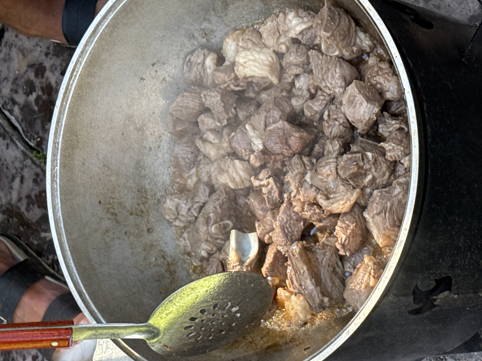 A close-up, overhead shot captures a large, black metal pot filled with simmering chunks of meat, likely beef, in a savory broth. The meat is a mix of brown and lighter shades, with some pieces showing a slightly gelatinous texture. A large metal ladle with a wooden handle, its bowl perforated with circular holes, rests in the pot, partially submerged in the liquid and nudging some of the meat. Wisps of steam rise from the pot, indicating active cooking. In the background, blurred legs of at least two individuals are visible, clad in dark sandals with dark straps, standing on a damp, uneven surface that appears to be cobblestone or rough pavement. The overall scene suggests an outdoor cooking event, possibly a traditional Kyrgyz meal preparation, given the context of Sary-Oy, Kyrgyzstan, a village known for its hospitality and traditional cuisine. The environment appears rustic and functional, emphasizing the cooking process.