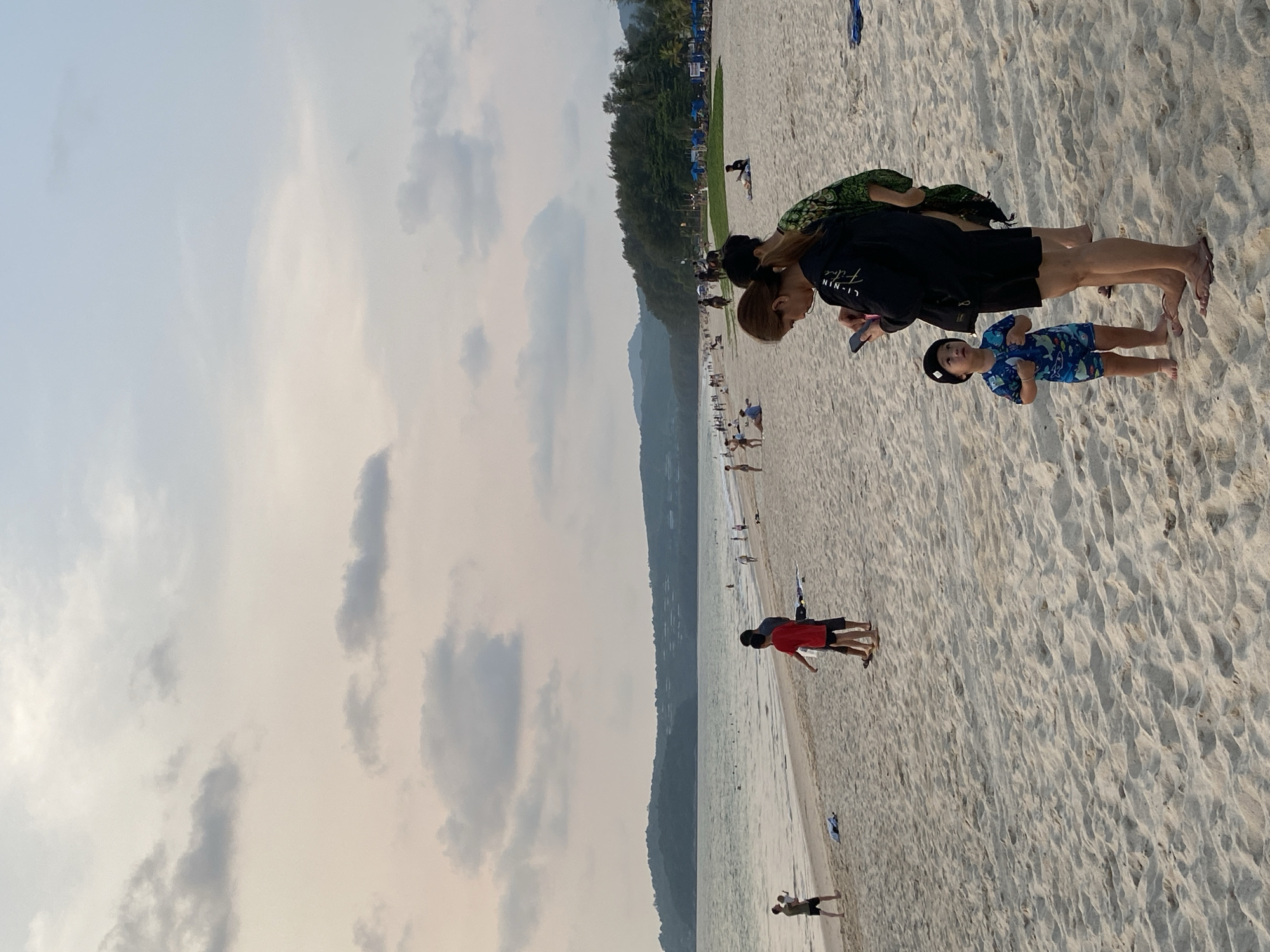 This image captures a broad, sandy beach in Choeng Thale, Thailand, under a soft, diffused light, suggesting late afternoon or early evening. The sky is partly cloudy with light grey and white clouds. The calm ocean water stretches along the left side of the frame, meeting a long, pale sand beach that extends into the distance. A line of lush green tropical trees borders the back of the beach, with distant mountains or hills rising behind them.
In the foreground, a woman with brown hair, wearing a black t-shirt with "LI-NIN" printed in white and dark shorts or a skirt, stands barefoot on the sand. A green patterned sarong or cover-up is draped over her shoulders. Beside her is a young child, dressed in a blue patterned swim outfit and a dark cap, also barefoot. Both are looking towards the left.
Further along the beach, numerous other people are visible enjoying the coastal environment. In the mid-ground, two individuals, one wearing a red t-shirt and dark shorts, are standing near the water's edge, with what appears to be a surfboard leaning on the sand nearby. Many smaller figures dot the sandy shore, some walking, others wading in the shallow water, indicating leisure activities. The overall scene conveys a relaxed and tranquil atmosphere at a popular beach destination.