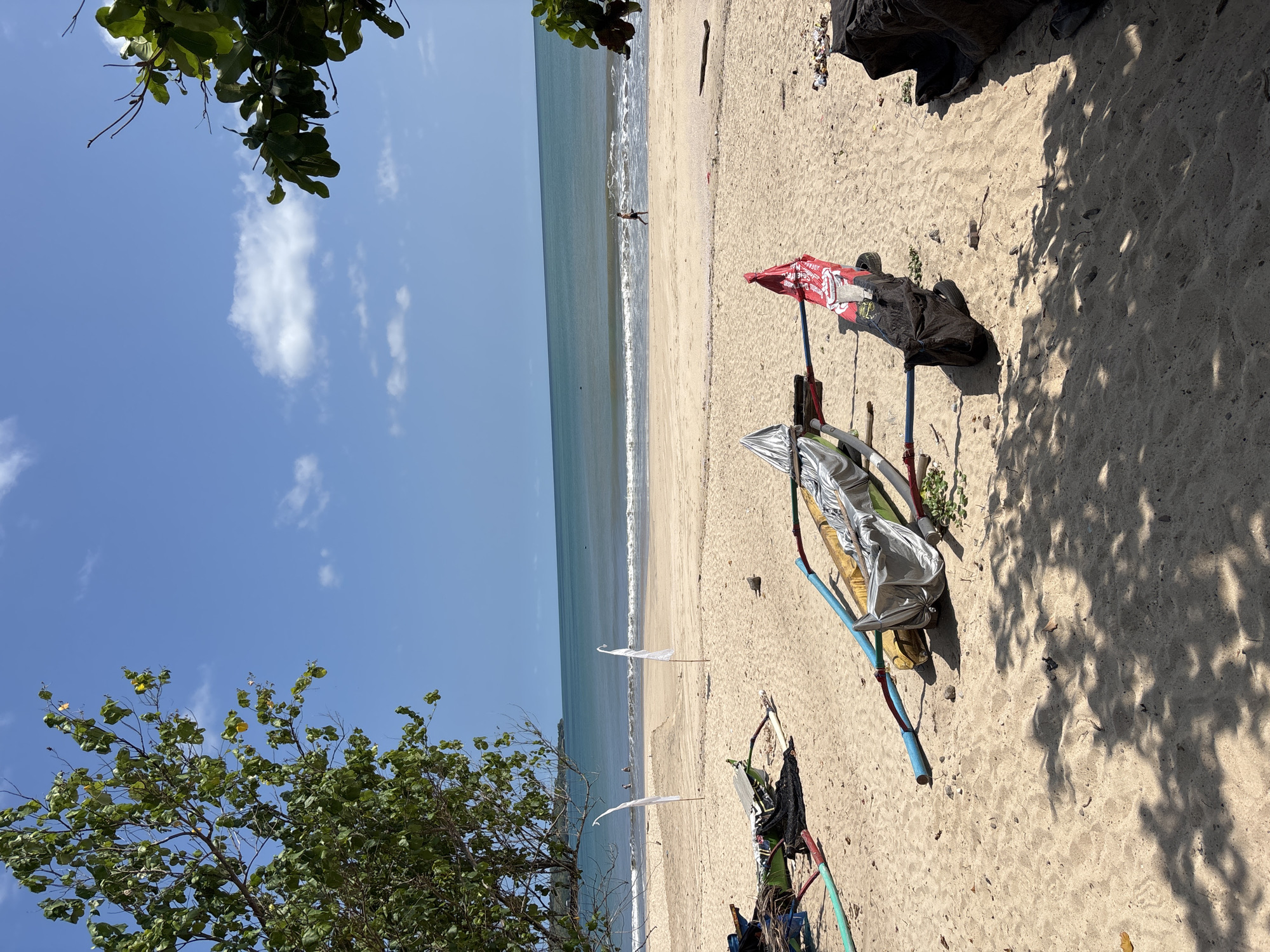 The image depicts a beach scene in Kuta, Indonesia, on a sunny day with scattered clouds. The shoreline is visible in the background. In the foreground, there are objects on the sand, including a wheeled cart covered with various items. The cart is partially shaded, with shadows cast by nearby trees. A red flag is attached to the cart. Also present are white flags further away from the cart. A person is visible in the distance, walking along the beach. Trees are visible on both the left and top of the frame.