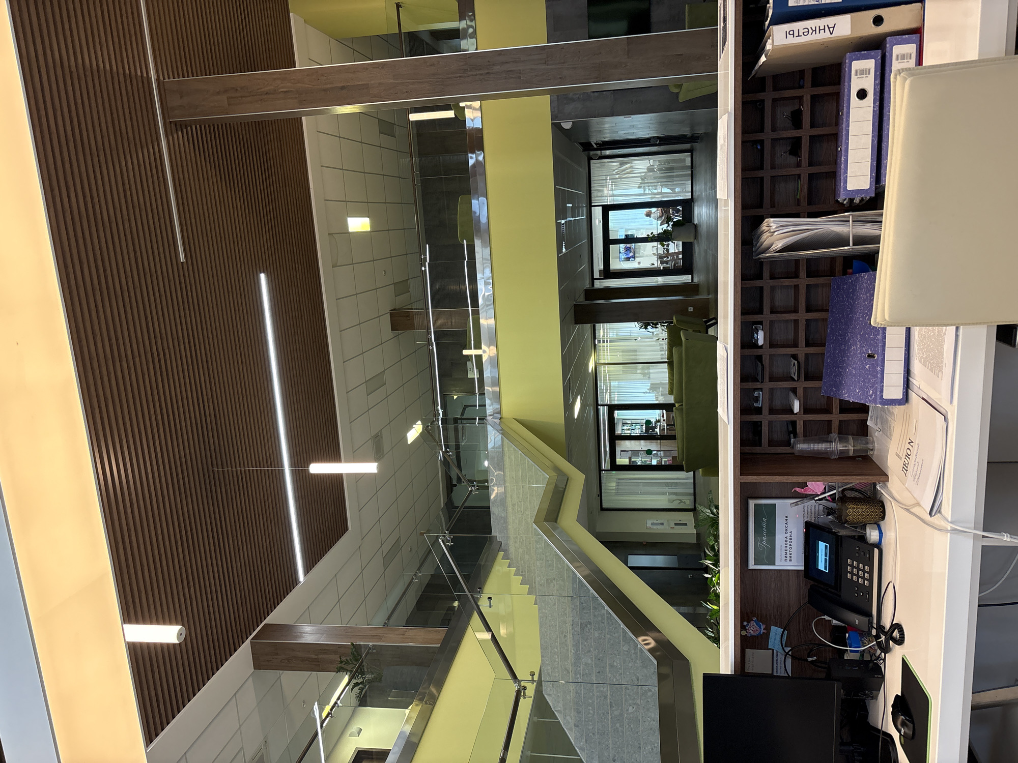 The image depicts the interior of a modern office building. The scene is dominated by a multi-level atrium with a striking wooden slatted wall and bright yellow accents. A contemporary staircase with glass railings winds through the space, connecting different floors.
On the right side of the image, a reception desk is visible. It is cluttered with office supplies, including a telephone, a computer monitor, a mouse, binders, and papers. A certificate with Cyrillic text is displayed on the wall behind the desk. There are also some decorative items and plants on the desk and shelves.
In the background, through glass doors, glimpses of other office spaces can be seen, with people working and sitting in seating areas. The overall impression is one of a clean, organized, and professional environment. The lighting suggests it is daytime. The location context is Kholmy, Russia.
Visible text includes "АНКЕТЫ" on a folder, and text on the certificate which appears to be a name and possibly an award. There is also text on some documents on the reception desk.
