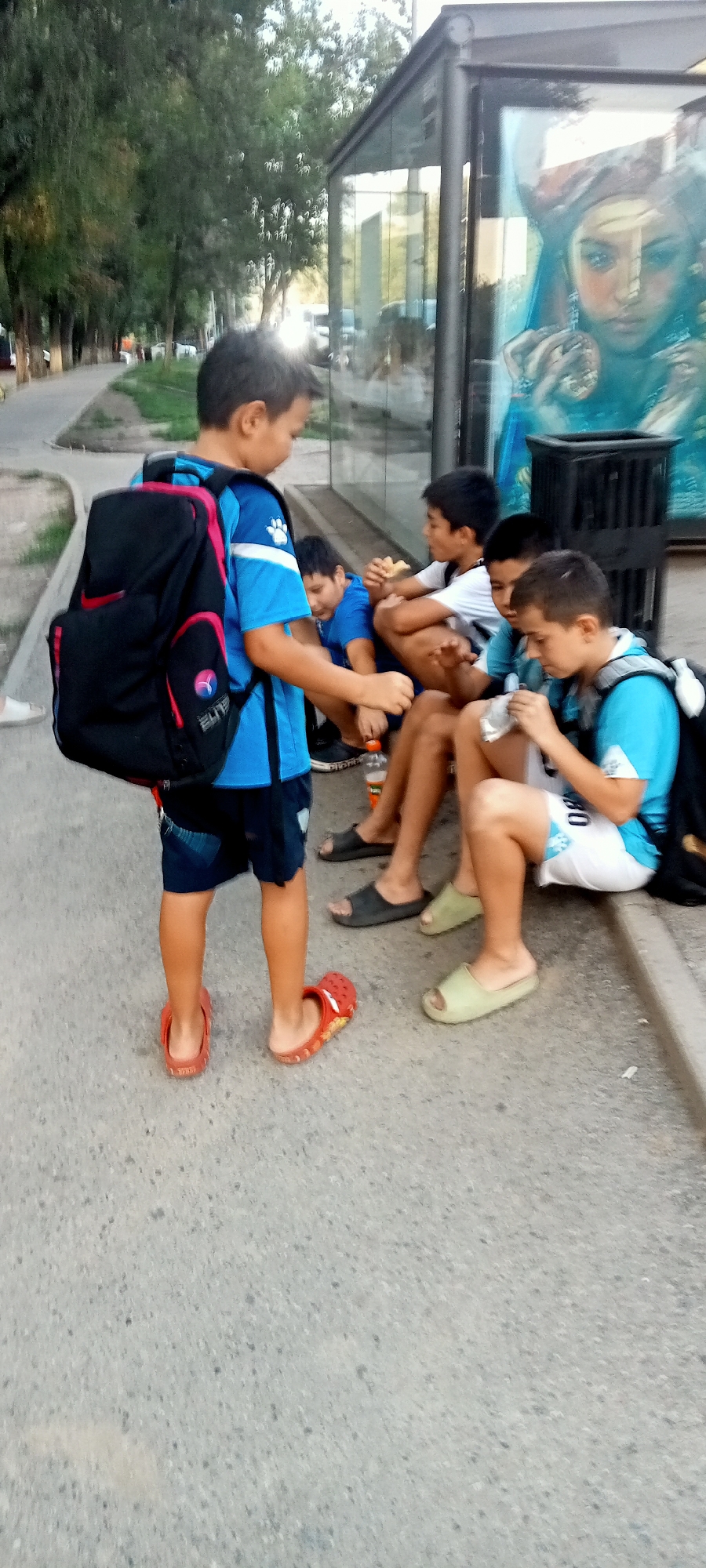 The image shows a group of boys near a bus stop with a mural on the glass. The boy in the foreground is wearing a blue t-shirt, dark shorts, red crocs, and a black backpack with pink accents. He is standing and facing towards the right side of the image. To his right, four other boys are sitting on the ground. One boy in a blue t-shirt is eating. Another boy in a white t-shirt is also eating. Two other boys in blue t-shirts are also present. They are sitting near a trash can. The bus stop appears to be located in Almaty, Kazakhstan, based on the architectural style and the mural which features an artistic representation that is consistent with urban art found in Almaty. The environment suggests an outdoor urban setting, likely a street or sidewalk area.