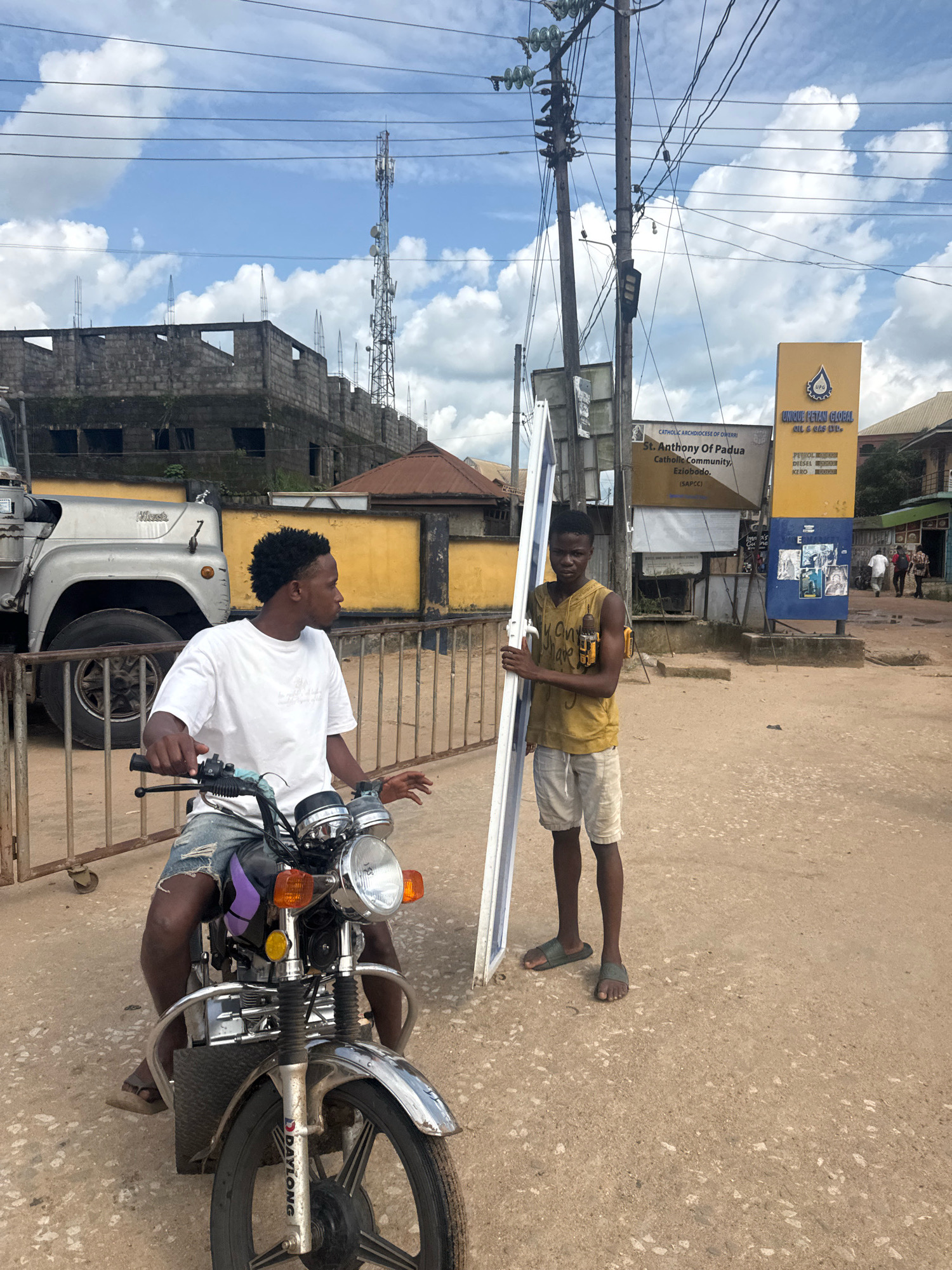 In Ihiagwa, Nigeria, on a dusty street, two young Black males are present. The first, seated on a silver motorcycle, wears a white t-shirt and distressed denim shorts. He grips the handlebars, looking to his right. The second male stands to the right of the motorcycle, holding a long, white rectangular object, possibly a window frame or solar panel, vertically. He wears a mustard-yellow sleeveless shirt and light-colored shorts, and has a yellow power drill attached to his right hip. Behind them, a large silver truck with "Mack" branding is parked. A yellow building with a dark roof is visible to the left, and further right, a tall yellow sign for "UNIQUE PETAN GLOBAL OIL & GAS LTD." stands beside a utility pole with numerous wires. The sky is partly cloudy.