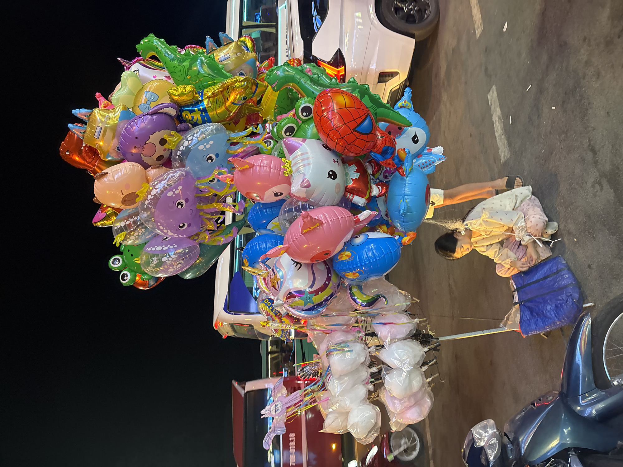 This media file captures a vibrant street scene at night in Nha Trang Ward, Vietnam. The primary subjects are a large cluster of colorful, character-shaped balloons, predominantly featuring sea creatures like octopuses and dolphins, alongside other popular characters like a dinosaur, a frog, and Spider-Man. Interspersed with the balloons are bags of cotton candy, some white and some pink, displayed on a cart.
A person, likely a vendor, is partially visible, sitting or lying down amidst the merchandise. They are wearing light-colored clothing and a blue skirt. Their posture suggests a moment of rest or waiting. The setting is a paved street, illuminated by artificial light, indicating it is nighttime. A white vehicle is parked nearby, and parts of a red vehicle or cart are visible in the lower portion of the image. A motorcycle is parked in the foreground on the right.
The overall mood is lively and whimsical due to the bright colors and playful shapes of the balloons, contrasting with the dark night sky. The scene depicts a typical street vending setup, common in many tourist areas, where such items are sold to passersby, especially during evening hours. The presence of various vehicles and the paved road suggest an urban or semi-urban environment. There is no visible text in the image.