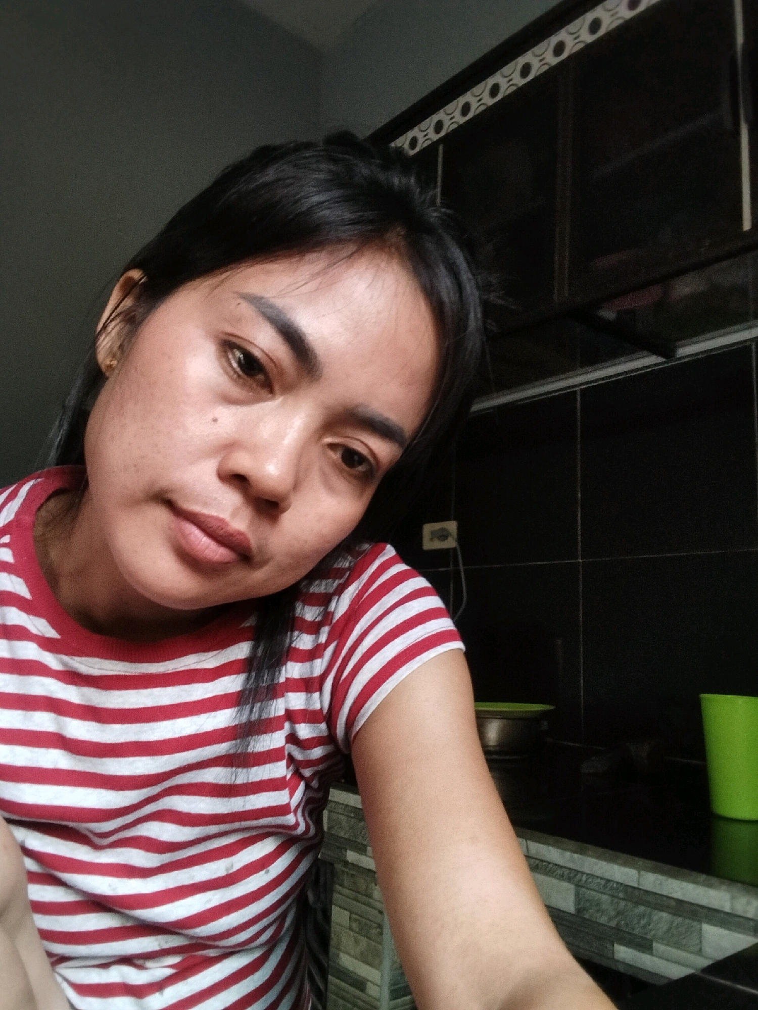 An Asian woman with dark hair and fair skin is pictured in a close-up, waist-up shot. She is wearing a red and white horizontally striped t-shirt. Her head is tilted slightly to her left, and she is looking towards the camera with a neutral expression. The background appears to be a kitchen area with dark cabinets and tiled walls. A green cup is visible on the right side of the frame. The lighting is somewhat dim.