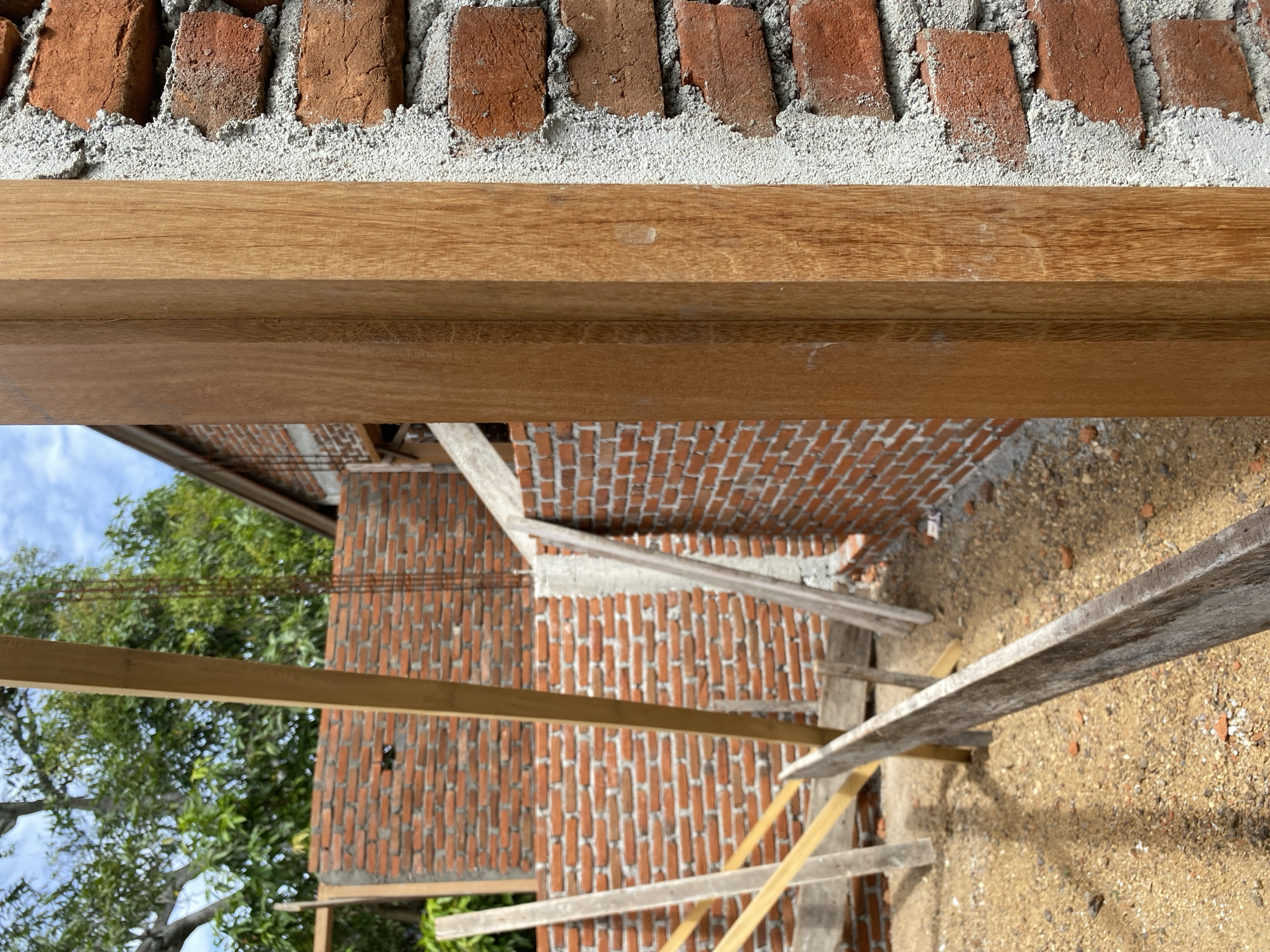 In the foreground of an outdoor construction site, a vertical wooden post stands to the right, adjacent to a brick wall. The brick wall, constructed with red bricks and white mortar, is partially completed. Behind the wooden post and brick wall, the construction continues with more brick walls, wooden beams, and scaffolding. The ground is covered in gravel and sand, with scattered debris. Trees and a cloudy blue sky are visible in the background, suggesting it is daytime. The image captures a scene of ongoing building work, likely a residential or commercial structure.