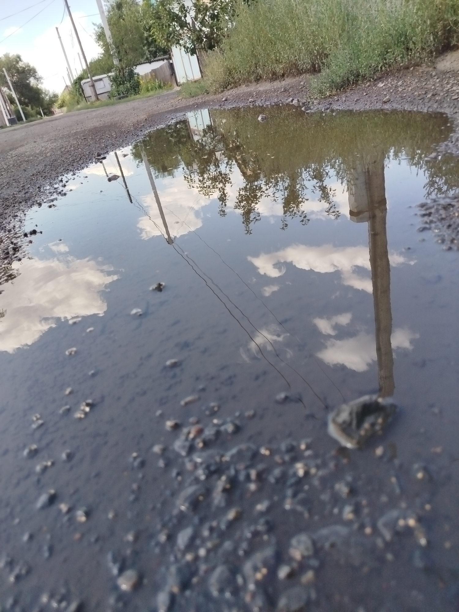 A shallow puddle of water reflects the sky and surrounding environment on a gravel road. In the reflection, a tall, slender pole, possibly a utility pole, stands prominently. Overhead power lines are visible, stretching across the frame, also reflected in the water. The road surface is uneven, with visible gravel and small stones. In the background, there are trees and what appears to be a fence and some structures, suggesting a semi-urban or rural setting. The overall impression is one of a simple, everyday scene on a clear day.