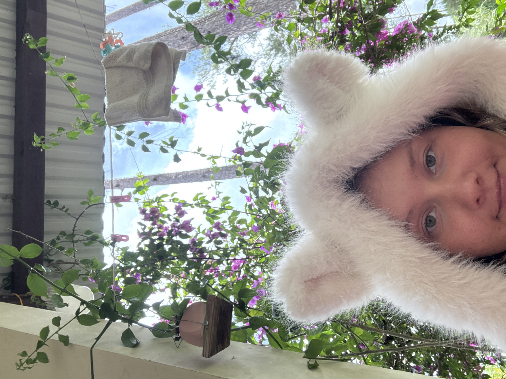 A person wearing a white, fluffy hooded garment with attached ears is positioned in the foreground, facing the camera. The individual's face is partially visible, with one eye looking towards the camera. The background is filled with lush green foliage and vibrant purple flowers, suggesting an outdoor environment. A clothesline with a beige towel and colorful clothespins is visible, suspended above the foliage. A pink planter with a plant is mounted on a wooden shelf to the left of the frame. The sky above is bright blue with scattered clouds, indicating daylight. The scene appears to be in a tropical or subtropical climate, possibly a residential area or garden in Đạ Cháy, Vietnam, characterized by abundant plant life and an outdoor living space.