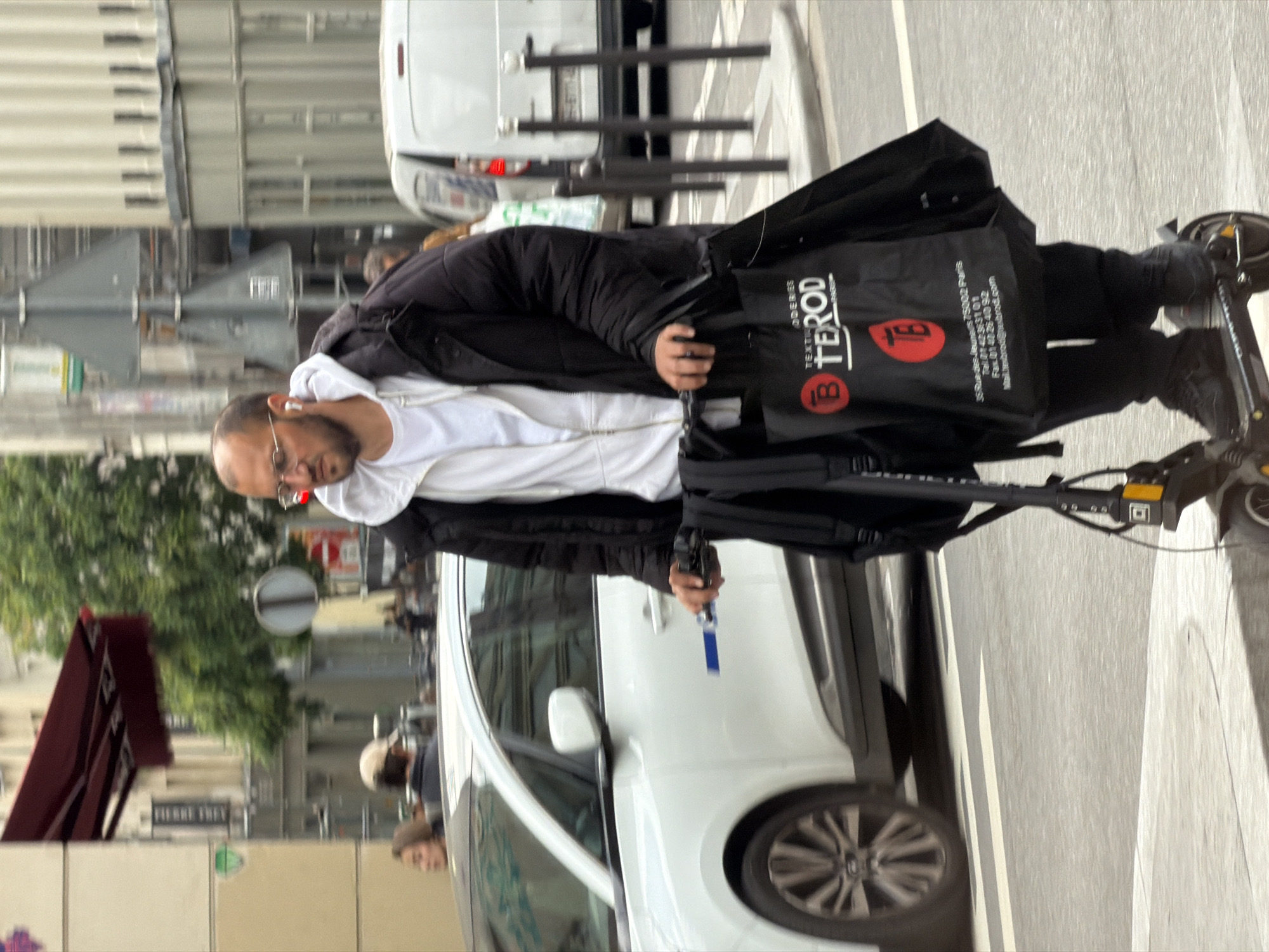 A man on an electric scooter is riding on a city street in Paris, France. He is wearing a white hooded sweatshirt, a black jacket, glasses, and has an earpiece in his left ear. He is holding a black bag with a red logo and the word "TEROD" printed on it. The scooter is traveling on a crosswalk in front of a white car. A Metro sign, labeled "PIERRE THIETRY," is visible in the background to the left of the car. Other pedestrians and vehicles can be seen nearby, including a white van. The scene is set in an urban environment with buildings and trees lining the street. The street appears to be paved with white stone and marked with white lines.