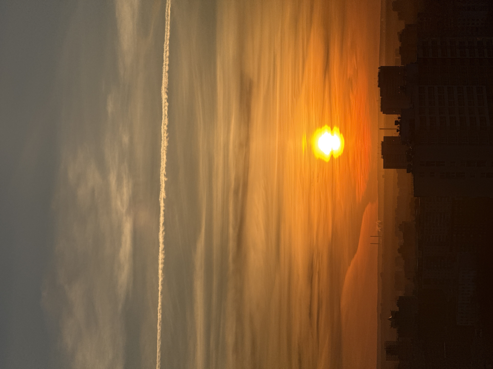 The image captures a dramatic sunrise or sunset over an urban landscape. The sky dominates the upper portion of the frame, showcasing a striking horizontal gradient from a muted grey-blue on the left to a vibrant, fiery orange and yellow closer to the horizon on the right. The sun, a brilliant and intense glowing orb with some visual flaring, is prominently positioned above the city silhouette, radiating warm light across the atmosphere. Streaks of wispy, elongated clouds span the middle section of the sky, catching the warm hues and creating a painterly effect. A distinct, straight white contrail (or vapor trail) stretches horizontally across the upper-left part of the sky, providing a sharp linear contrast against the softer cloud formations.
Below the illuminated sky, the silhouette of a city skyline is visible. On the far right, dark, tall buildings rise against the bright backdrop, their architectural details largely obscured by shadow. Further towards the center and left, fainter silhouettes of distant structures, including what appear to be industrial chimneys or towers, can be discerned. A dark, calm body of water, likely a river or large lake, extends across the mid-ground, subtly reflecting some of the sky's light as it meets the distant landmass. The scene evokes a sense of serenity and natural grandeur, clearly depicting either dawn or dusk. There is no visible text within the image.