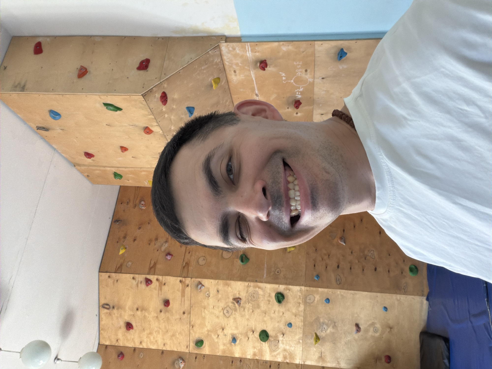 A man wearing a white shirt smiles in front of a climbing wall. The wall is made of wood panels with various colored climbing holds attached. There is mathematical notation visible on the wall, potentially related to climbing routes or problem-solving. The environment suggests an indoor climbing gym. I cannot confirm the location in the city of Izhevsk, Russia.