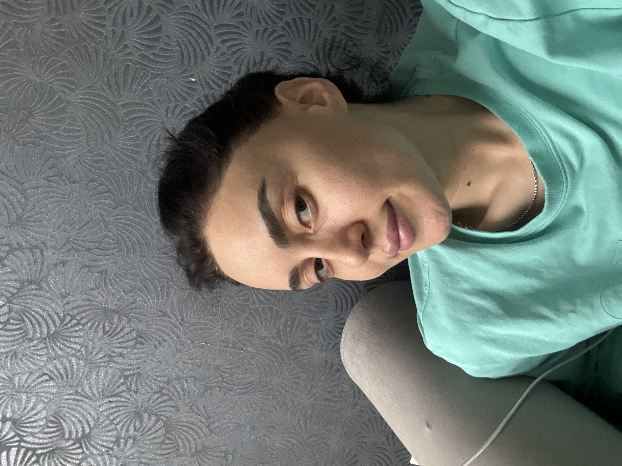 The photograph captures a person, identified as female, lying on their back in an indoor setting. The image is oriented such that her head is on the left and her body extends to the right. She has dark hair, pronounced dark eyebrows, and looks directly at the camera with a neutral to slightly smirking expression. She is wearing a light turquoise-colored t-shirt, which reveals a silver chain around her neck. Her lower body, partly visible, is clad in what appears to be light grey casual wear, possibly shorts or pants. A thin white cable runs across the lower right portion of the frame. The background consists of a dark grey surface featuring a textured, repeating pattern of stylized fan or feather shapes, suggesting a decorative blanket, rug, or upholstered furniture. The lighting is soft and even, typical of an indoor environment. The overall mood is relaxed and informal. No visible text is present in the image. The user-provided context indicates the location is Tbilisi, Georgia.