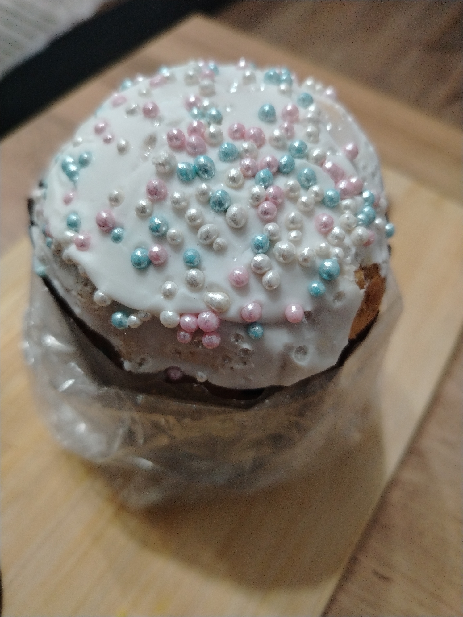 A close-up shot shows a Kulich, a traditional Russian Easter bread, decorated with white icing and colorful sprinkles. It's wrapped in clear plastic wrap and rests on a light-colored wooden surface. The sprinkles are predominantly white, pink, and light blue.
There is no information to indicate this image was taken in the city of Izhevsk, Russia, or any other location. There is no information available regarding people, actions, or interactions.