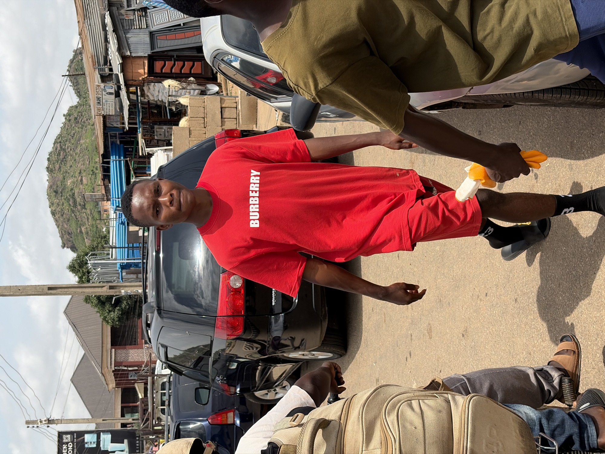 In the city of Gura Top, Nigeria, on a sunlit day, a young man walks forward wearing a red t-shirt with "BURBERRY" printed in white on it, and red shorts. He is also wearing black socks with white lettering and black sandals. Next to him, a person wearing a light brown t-shirt and blue pants walks to the right. To the left of the young man, another person is seen from the waist down, wearing a beige backpack and dark jeans. In the background, several vehicles are parked, including a black Toyota minivan. The scene is set along a paved road lined with buildings, some featuring corrugated metal roofs and others with tiled roofs. A hill rises in the distance under a hazy sky.