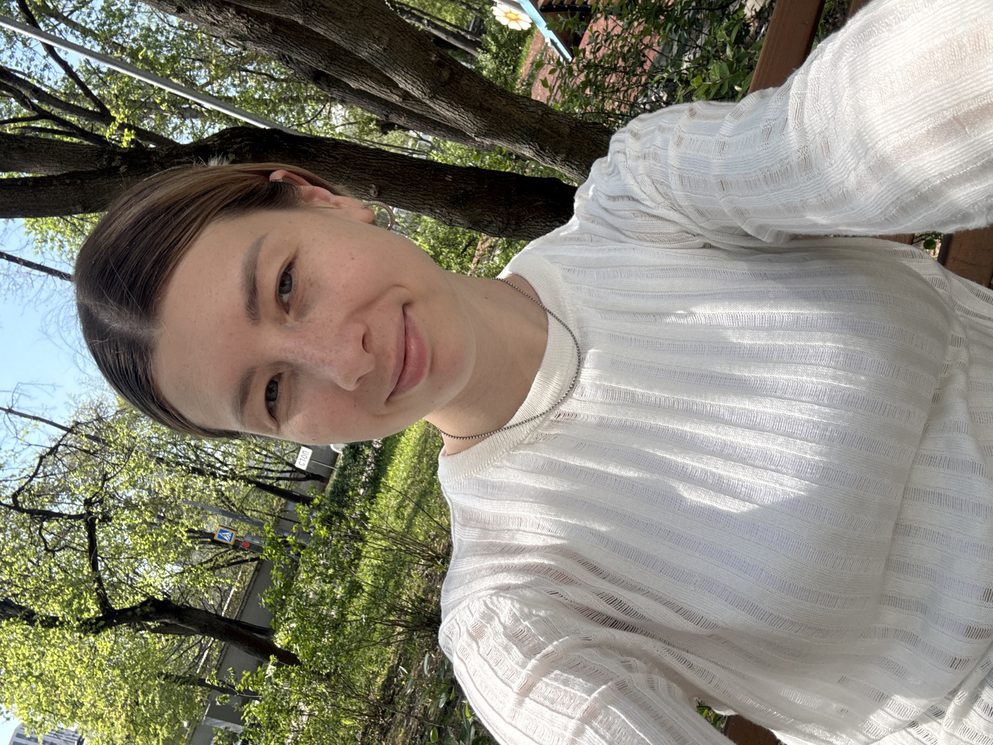 A young woman is posing for a selfie outdoors. She has light brown hair and is wearing a white, semi-sheer shirt with long sleeves and a delicate silver necklace. Her expression is a slight smile, and she is looking directly at the camera.
The background shows an outdoor setting with trees and greenery, suggesting a park or garden. A blue sky is partially visible through the trees. A street sign is seen with the Russian inscription 'СТОП'. A yellow street sign can be seen on the left side.
Based on visual cues, it is possible that the location of this photograph is in Moscow, Russia.