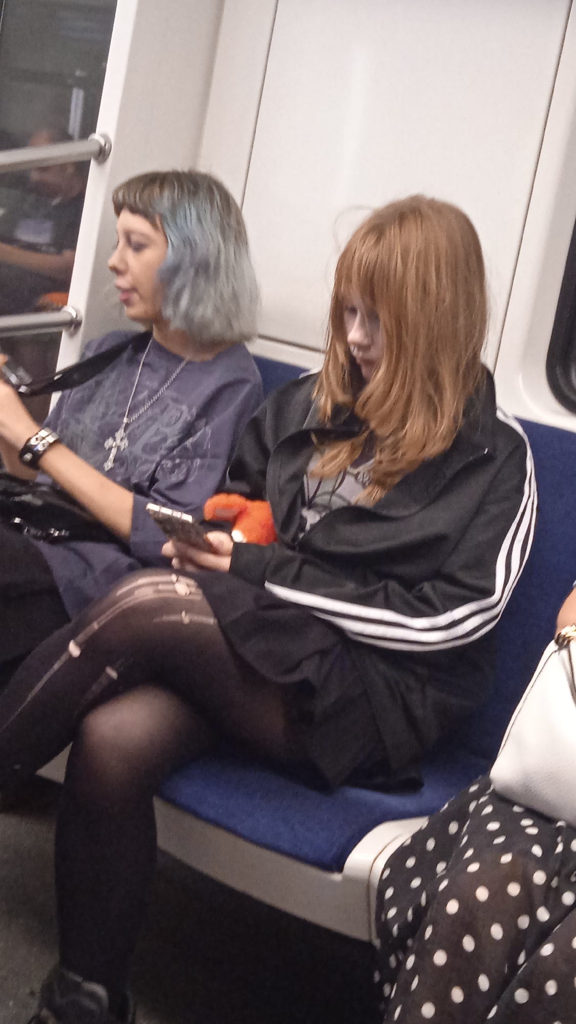 Two young women are seated side-by-side on a public transit vehicle, likely a train, as indicated by the interior design and the presence of windows and seating typical of such transportation. The woman on the left has light blue-grey, short, blunt-cut hair and is wearing a dark blue, long-sleeved shirt with a silver cross pendant. She is holding a smartphone in her right hand and has a black bracelet on her left wrist. The woman on the right has long, reddish-brown hair and is wearing a black jacket with white stripes down the sleeves over a grey t-shirt, and dark, possibly ripped, leggings. She is holding a smartphone in her right hand and has an orange stuffed toy in her left hand. Both women appear to be looking at their phones. The seats are blue, and the interior of the vehicle is light-colored. A portion of another person's arm and a white purse are visible on the far right of the image. The overall lighting suggests an indoor setting.