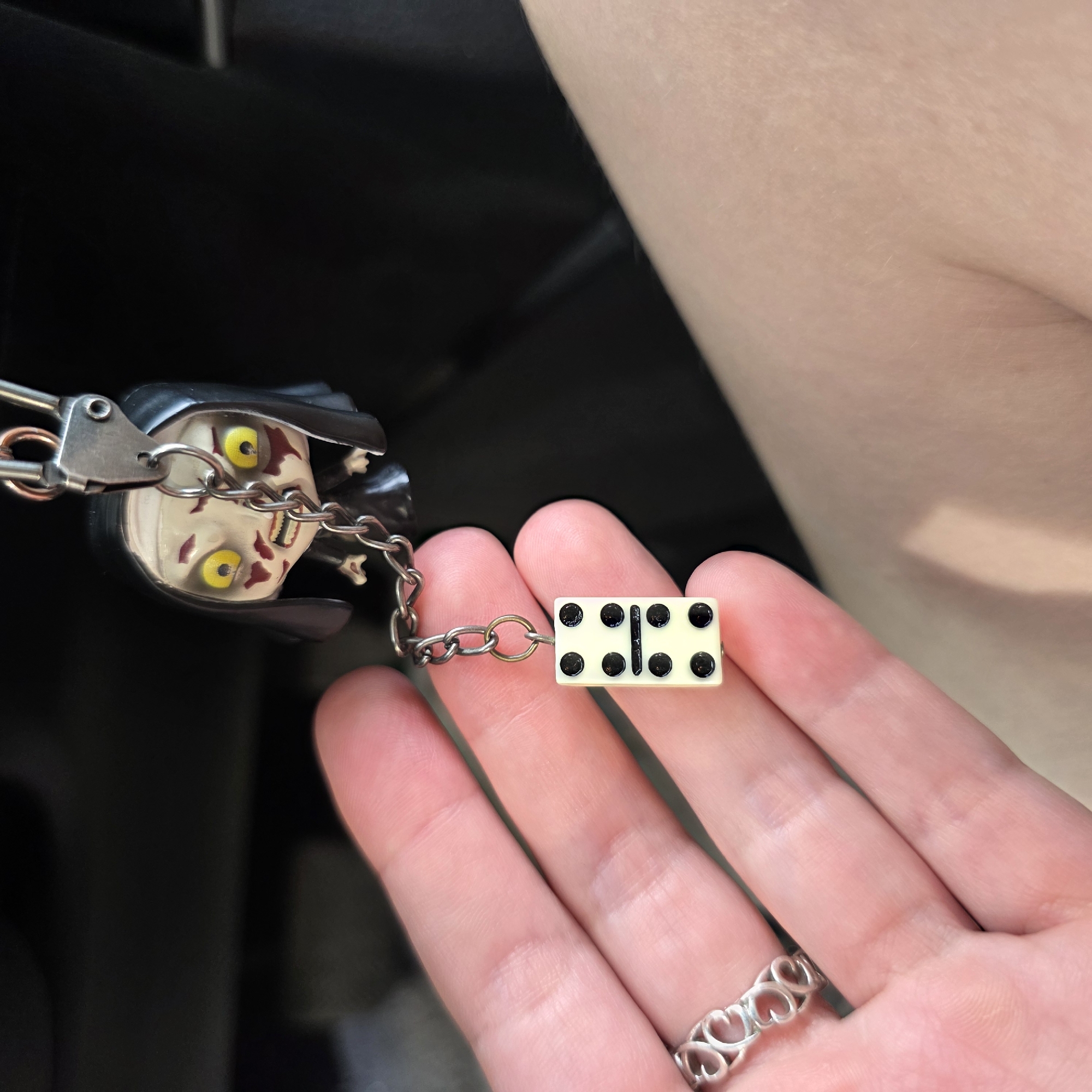 A hand with fair skin and a silver heart-shaped ring on the index finger holds a keychain. The keychain features a miniature doll with a white face, yellow eyes with red markings, and a black habit, resembling a demonic nun. Attached to the doll by a chain is a white domino charm with black pips. The domino is oriented vertically, with the top section displaying six pips and the bottom section displaying four pips, separated by a horizontal line. The background is dark and out of focus, suggesting an indoor setting such as a car interior.