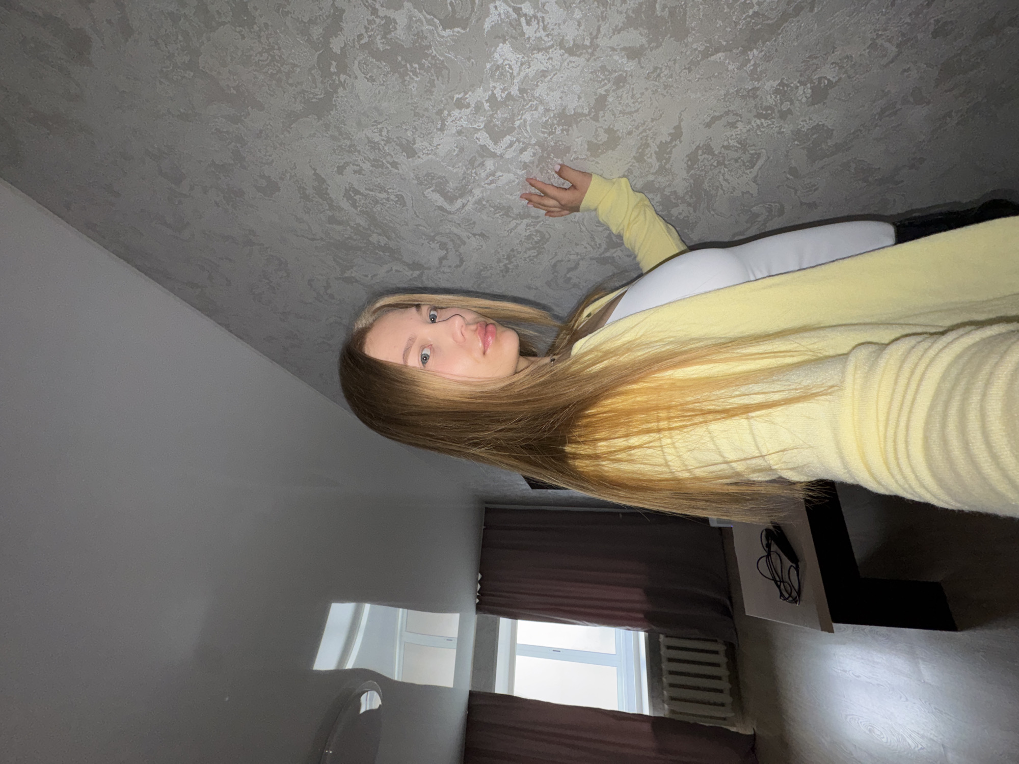 A young woman with long blonde hair, wearing a yellow cardigan over a white top, is pictured in what appears to be an indoor setting. The ceiling has a textured, grey finish. A window with dark curtains is visible in the background. Part of a white table with black accessories is on the right of the frame. Based on the window, curtains, and furniture, the location is potentially in a residential area in Chaikovsky, Russia.