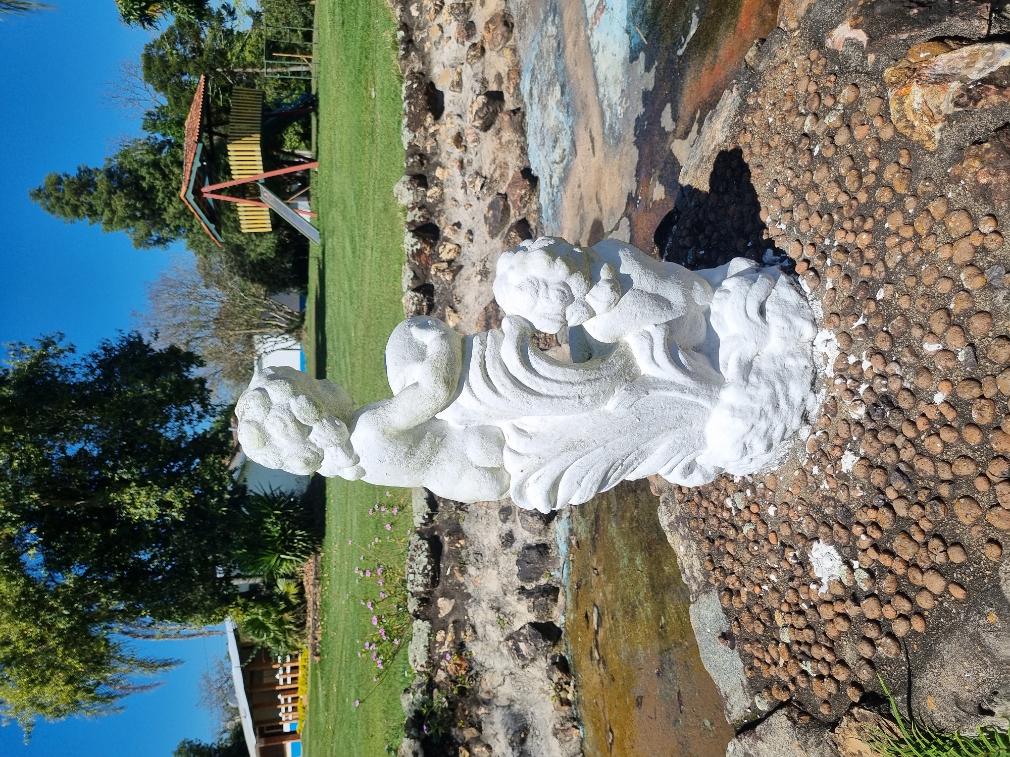 The image depicts a white stone statue of two cherubs in the foreground, situated on a circular stone platform covered with small, rounded brown pebbles. The statue is positioned next to a shallow body of water with rocky edges. Behind the statue, a well-maintained green lawn stretches out, dotted with small pink flowers. In the mid-ground, a house with a blue and white facade and a covered patio is visible on the left. On the right, a wooden playground structure with a yellow slide and a red frame stands on the grass. Tall green trees and a clear blue sky dominate the background, suggesting an outdoor setting. The scene appears to be located in a park or a residential area with ample green space, possibly within Laranjal Paulista, Brazil, given the context. There are no people or animals visible in the image.