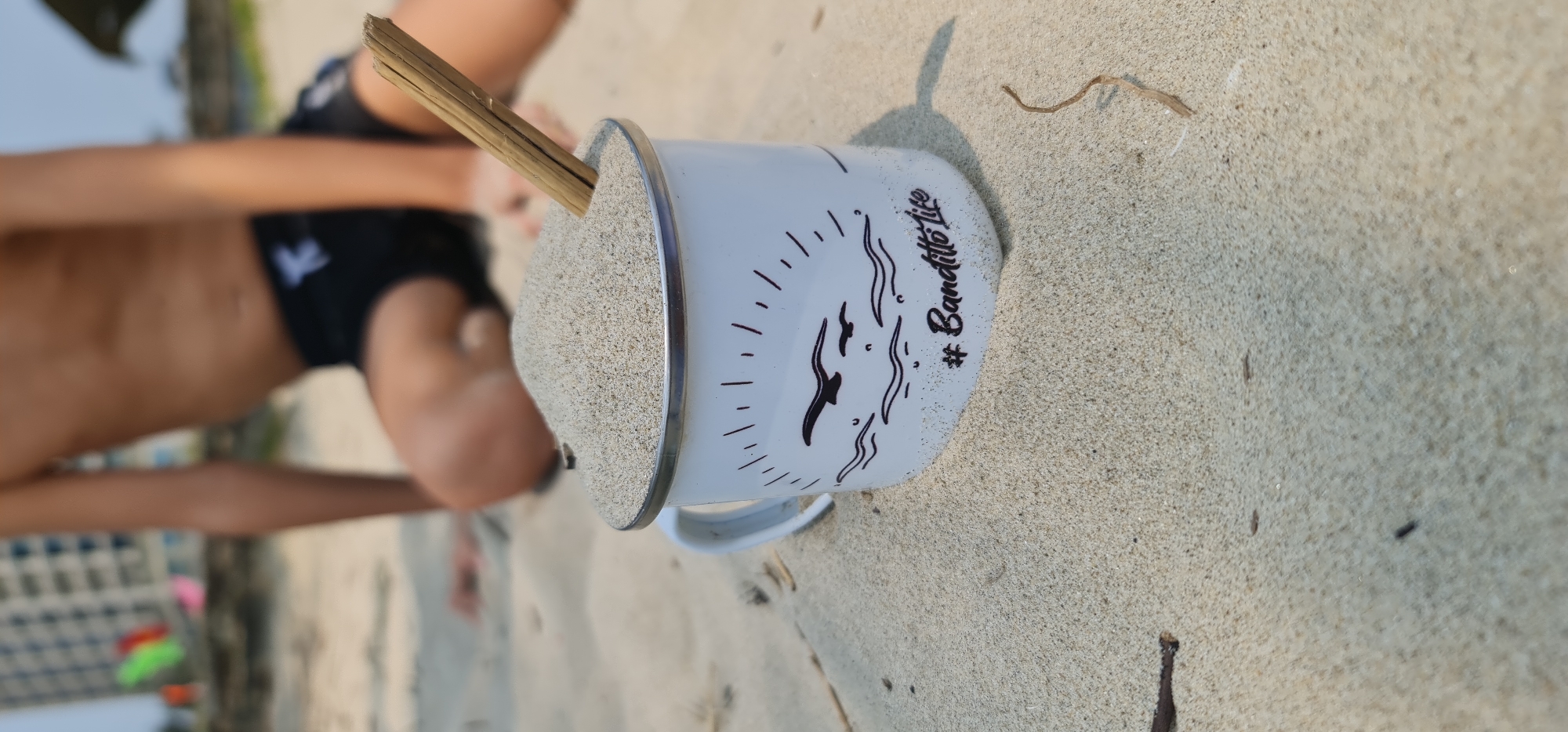 A low-angle, eye-level shot depicts a sand-filled mug on a beach, likely in Đà Nẵng, Vietnam, given the coastal setting and distant building architecture. The white mug with a silver rim features a minimalist design with black lines and waves, along with the hashtag "#Banditto Life" written on it. A small, wooden stick protrudes from the sand within the mug. A child's torso and arms are visible in the background to the left, suggesting they are sitting or lying on the beach. The sandy beach has scattered small pieces of debris and a few dark specks. In the far background, across the horizon, blurred buildings are visible, indicating a city location near the beach. The lighting suggests it is daytime, with a clear sky partially visible at the top left of the image.