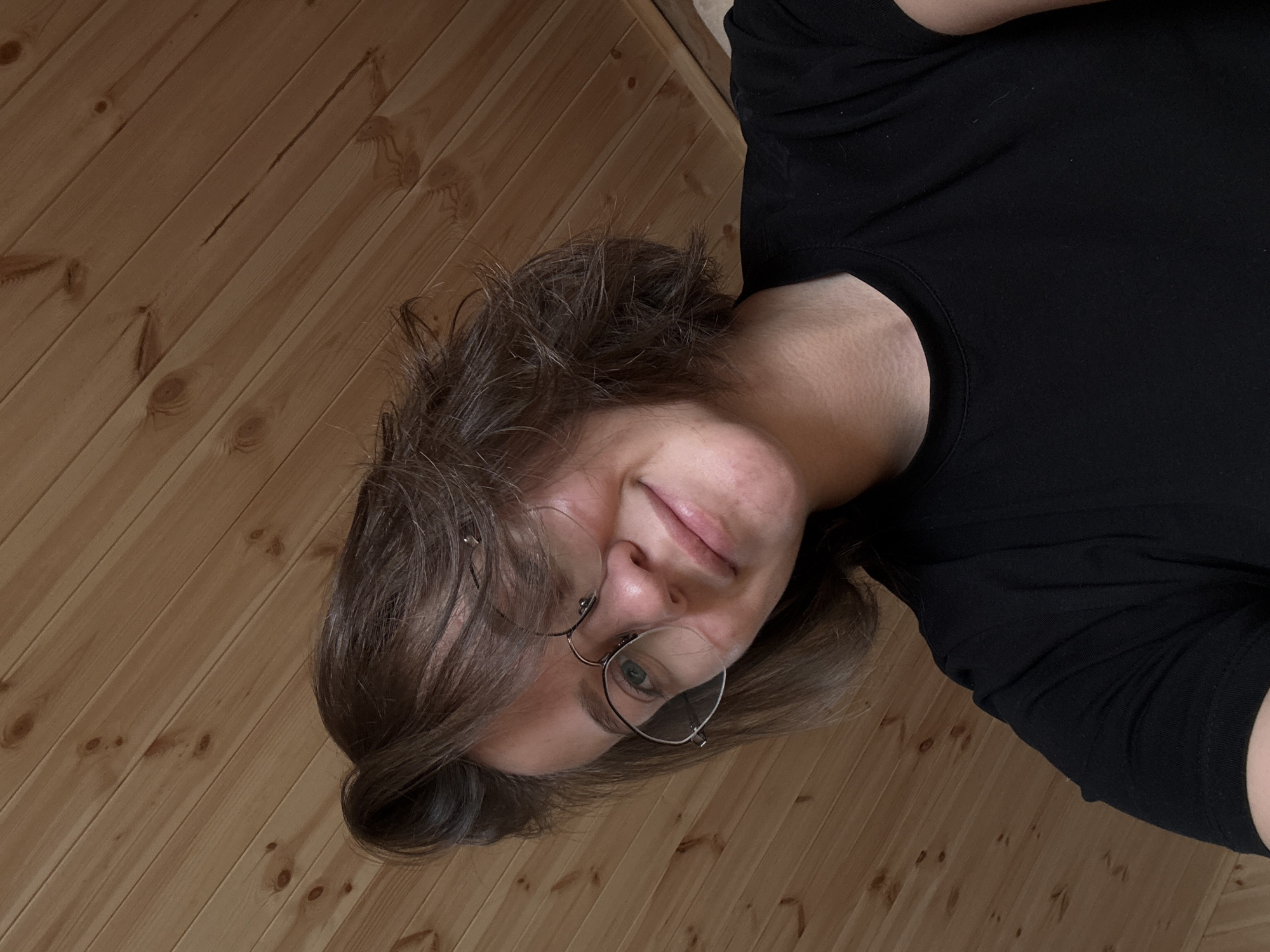 A young adult female is depicted in a close-up, upward-angled self-portrait. She has brown, wavy, bob-length hair, which is styled to her left, partially obscuring her right forehead and the top of her round, thin wire-rimmed spectacles. Her eyes appear light blue or green, and a subtle, closed-mouth smile is visible on her face as her gaze is directed upwards towards the camera. She is wearing a plain black, short-sleeved, crew-neck t-shirt. The background consists of light-toned wooden planks, oriented diagonally from the upper-left to the lower-right of the frame. The wood displays distinct grain patterns and scattered dark circular knots. The scene is evenly illuminated with soft lighting, characteristic of an indoor environment. This image is situated in the city of Lenino, Russia.