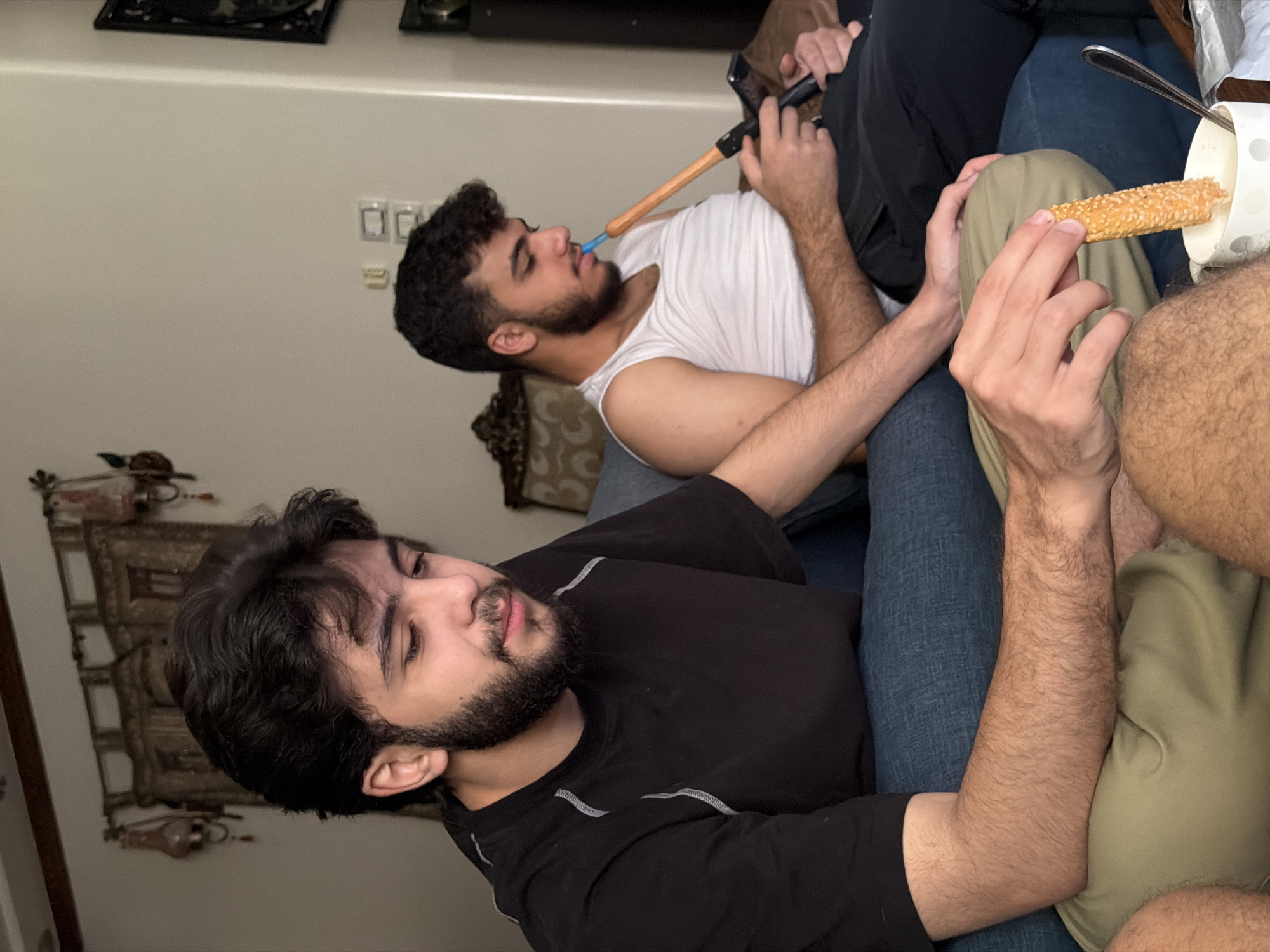 The image shows two young men indoors, likely in a residential setting in the Sarouja Municipality, Syria. The man in the foreground, wearing a black t-shirt, is seated and holding a long, bread-like snack, dipping it into a white mug with a polka-dot pattern. His gaze is directed downwards. To his right, another man in a white tank top is seated and holding a phone in his left hand while he uses a hookah with his right hand, inhaling from its pipe. The background suggests an interior space with decorative curtains, lamps, and light switches on the wall. The lighting is dim, casting soft shadows.