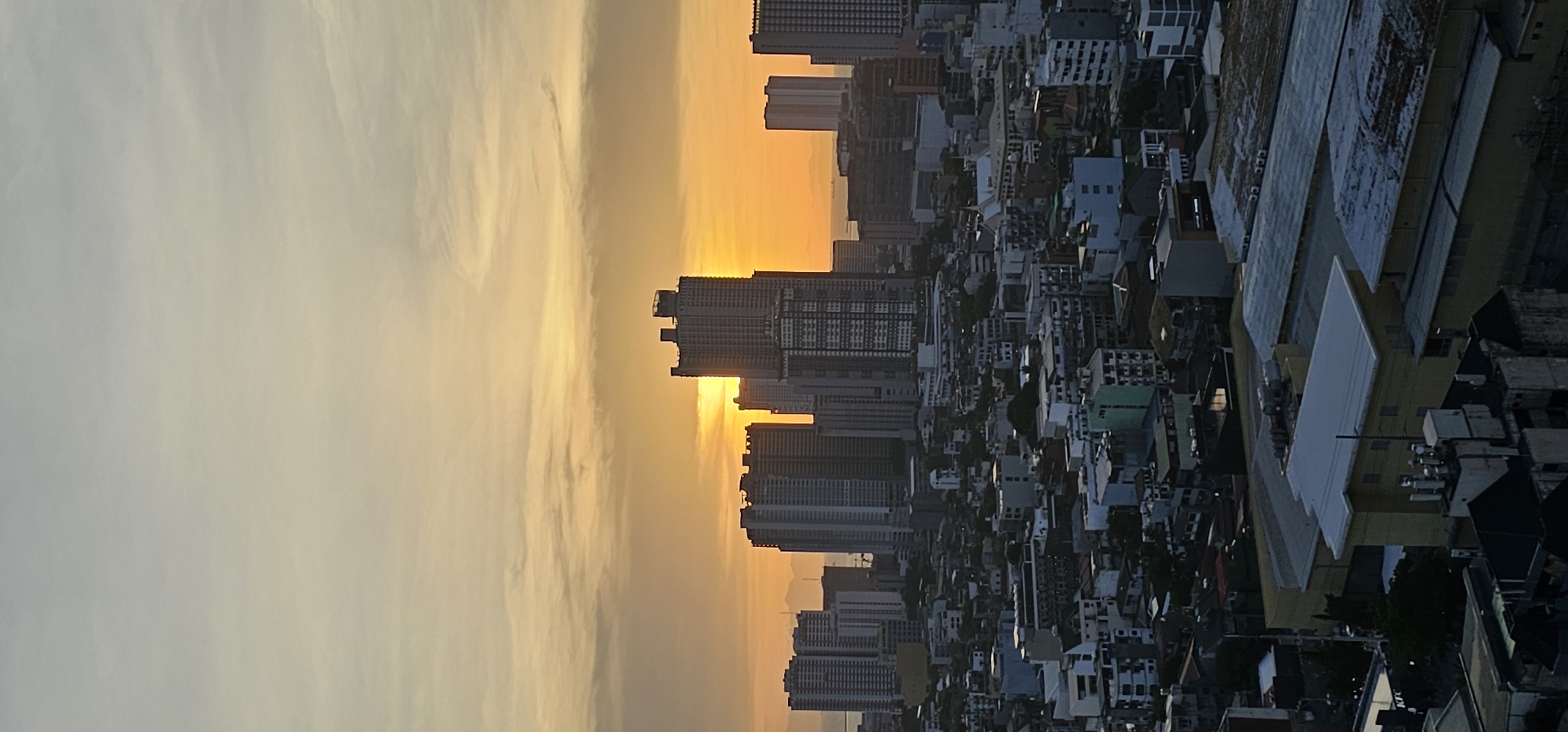 A panoramic view of the Makati cityscape in the Philippines is captured during sunset. The sky is a gradient of soft yellow and orange hues, with wispy clouds adding texture. Prominently featured are several high-rise buildings, some with distinctive architectural designs, silhouetted against the bright sky. The setting sun is partially obscured by one of the central skyscrapers, casting a warm glow over the scene. Below the towering structures, a dense urban landscape of lower buildings, rooftops, and streets is visible, showcasing the city's intricate layout. The image conveys a sense of the bustling urban environment of Makati, bathed in the dramatic light of dusk.