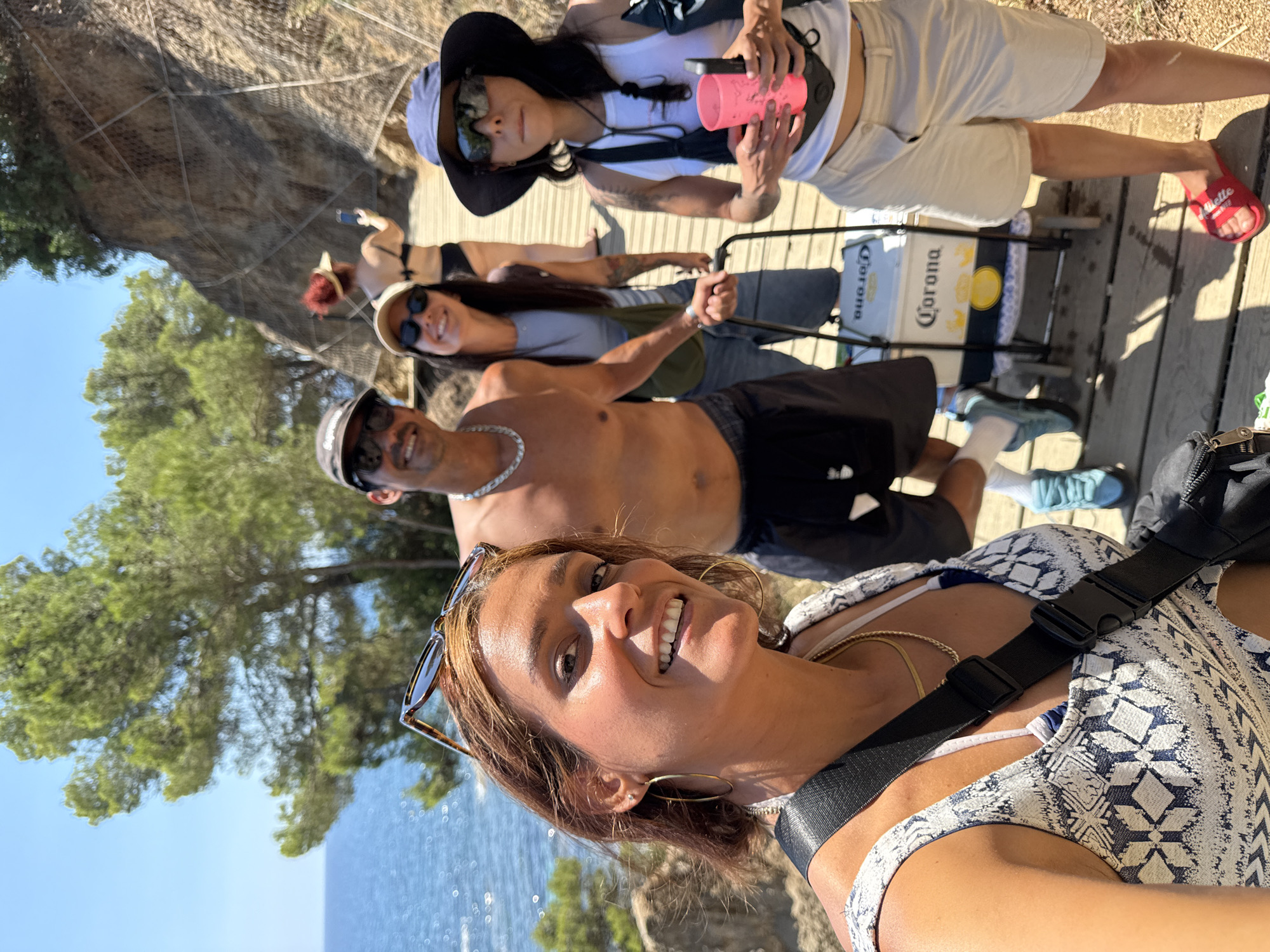 The image depicts a group of five people on a wooden boardwalk along a coastal path in Castell d'Aro, Platja d'Aro i s'Agaró, Spain. The sea is visible to the left, and a rocky cliff face with vegetation rises to the right. The boardwalk appears to be part of a scenic trail.
In the foreground, a woman with brown hair smiles at the camera, wearing a patterned top and a gold necklace. She has hoop earrings and sunglasses perched on her head. A black strap from a bag is visible across her chest.
To her right, a shirtless man with a chain necklace and sunglasses smiles, wearing dark shorts. Behind him, a woman in a light-colored top and a green crossbody bag also smiles. She wears a cap and sunglasses.
Further back and to the right, a woman in a white tank top, beige shorts, and a dark sun hat is holding a pink cup and a dark object. She wears sunglasses. To her left and slightly behind, another person with red hair is visible in a black bikini and a yellow visor, holding a phone.
A rolling cooler with a "Corona" logo is positioned between the second and fourth individuals. The environment suggests a sunny day, likely during a holiday or recreational outing.