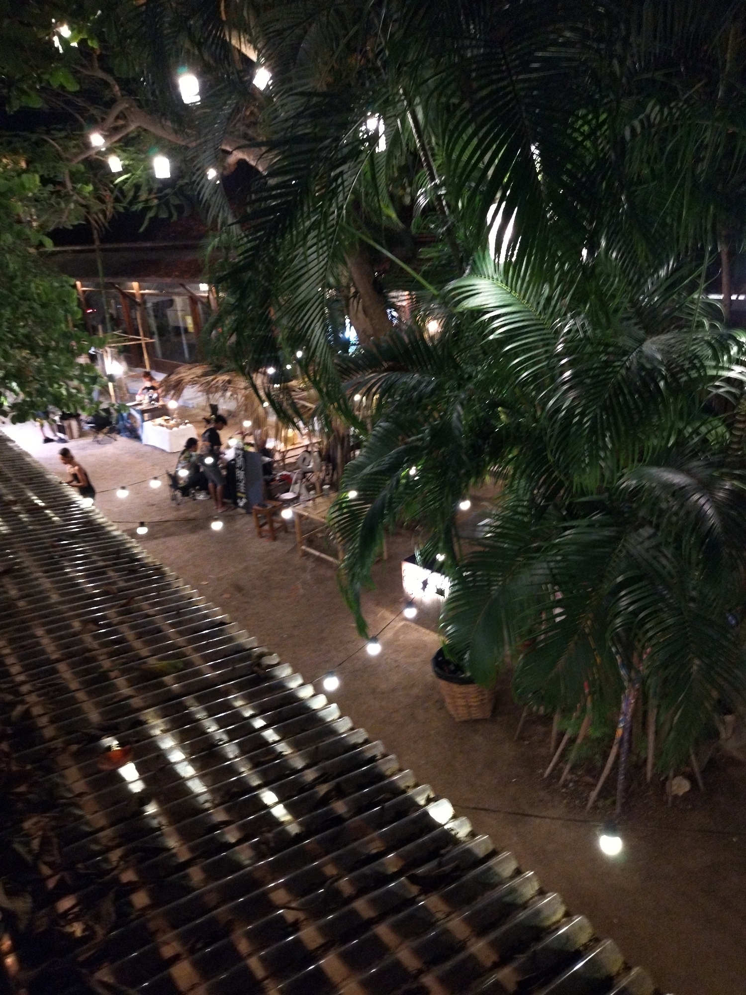 The image shows an outdoor market scene in ตำบลเกาะพะงัน, Thailand, at night. Several people are gathered at different stalls, appearing to browse or interact with vendors. String lights are hung across the space, illuminating the market area and creating a festive atmosphere. A corrugated metal roof dominates the foreground, with lights reflecting off its surface. In the background, lush greenery including palm trees is visible, adding to the tropical ambience.
