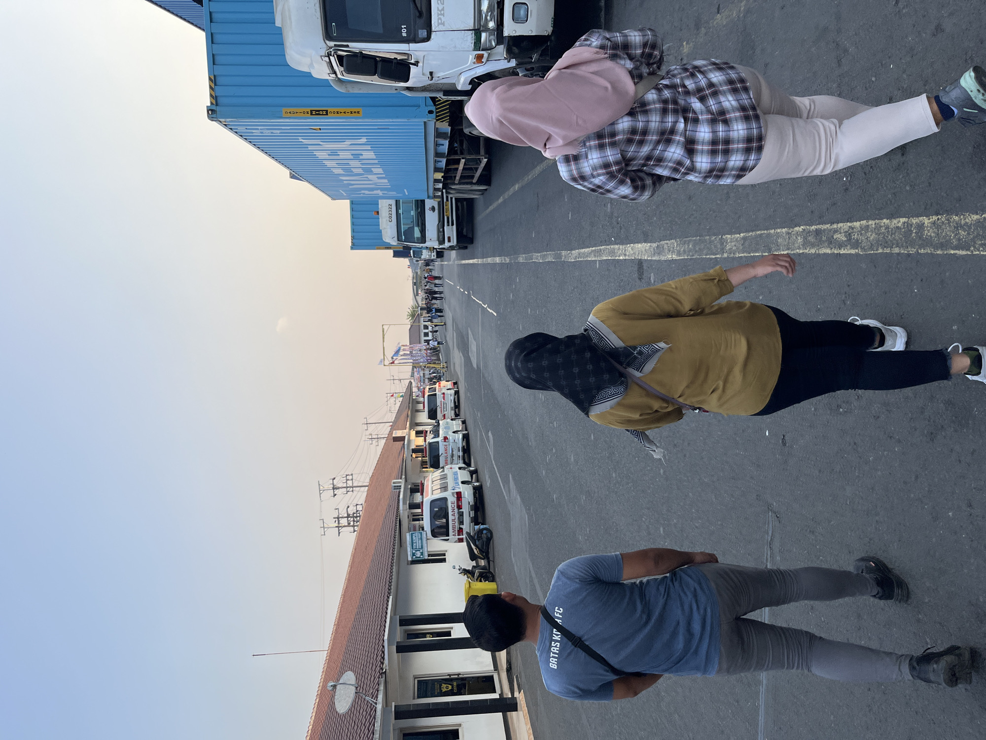 The image shows a street scene at what appears to be a port or industrial area. Three people are walking away from the camera down the middle of the road. The person in the foreground on the left is wearing a blue t-shirt with "BATAS KOTA FC" written on the back, grey pants, and black shoes. To their right, a person in a mustard-colored shirt and black pants with a black and white headscarf is walking. On the far right, a person in a plaid shirt and light-colored pants with a pink headscarf is walking.
The environment includes a long, low building with a tiled roof on the left, possibly an administration building, with several white vans, some marked "AMBULANCE," parked in front of it. Further down the road, large blue shipping containers are visible on the backs of trucks, with one white truck in the foreground. The sky is a pale blue, suggesting it is either early morning or late afternoon. The location is identified as the city of Pelang, Indonesia.