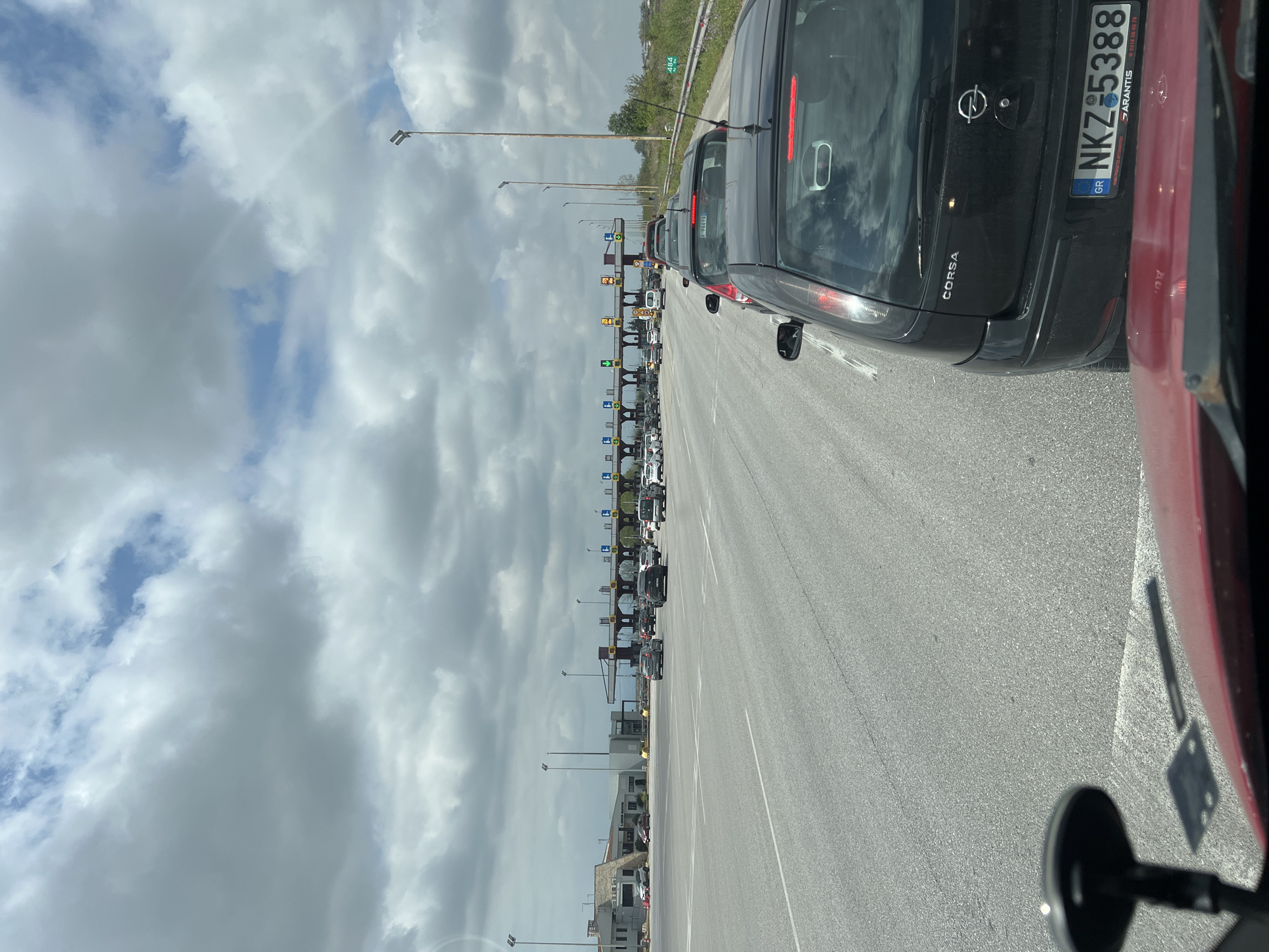 The image shows a long line of cars waiting at a toll booth. The license plate on the black Opel Corsa in front reads "NKZ 5388" and indicates that the car is registered in Greece. The location is in the Municipal Unit of Axios, Greece, as this is where the license plate is registered. The sky is cloudy, and there are street lights along the road.