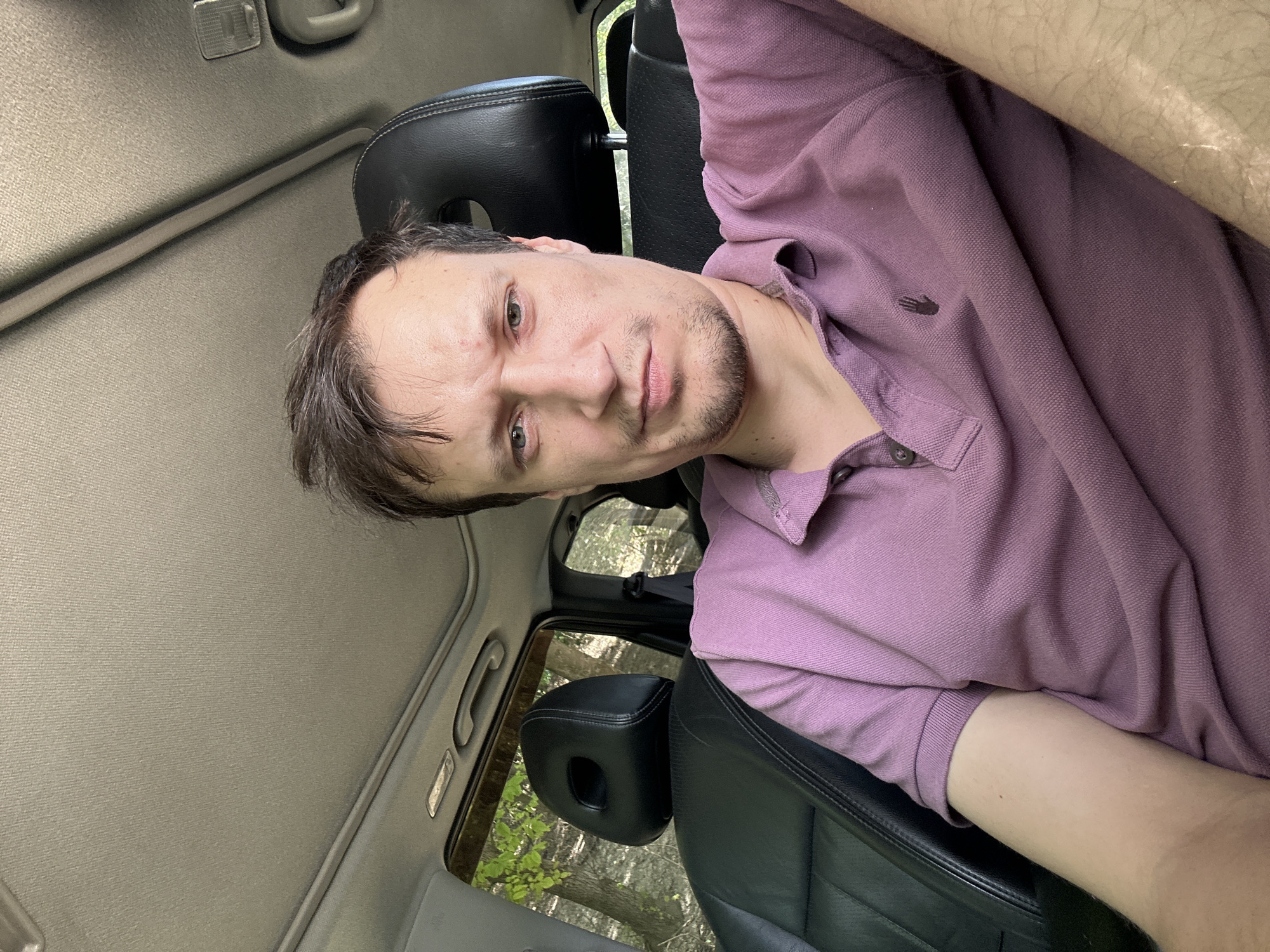 A man with short, dark hair and light-colored eyes is seated in the driver's seat of a car. He is wearing a purple polo shirt. The interior of the car is visible, including black leather seats and the car's headliner. Through the windows, trees and foliage can be seen, suggesting an outdoor, possibly wooded, location. The lighting indicates it is daytime. The image appears to be a selfie taken from the man's perspective. The location is the city of Makiv, Ukraine.