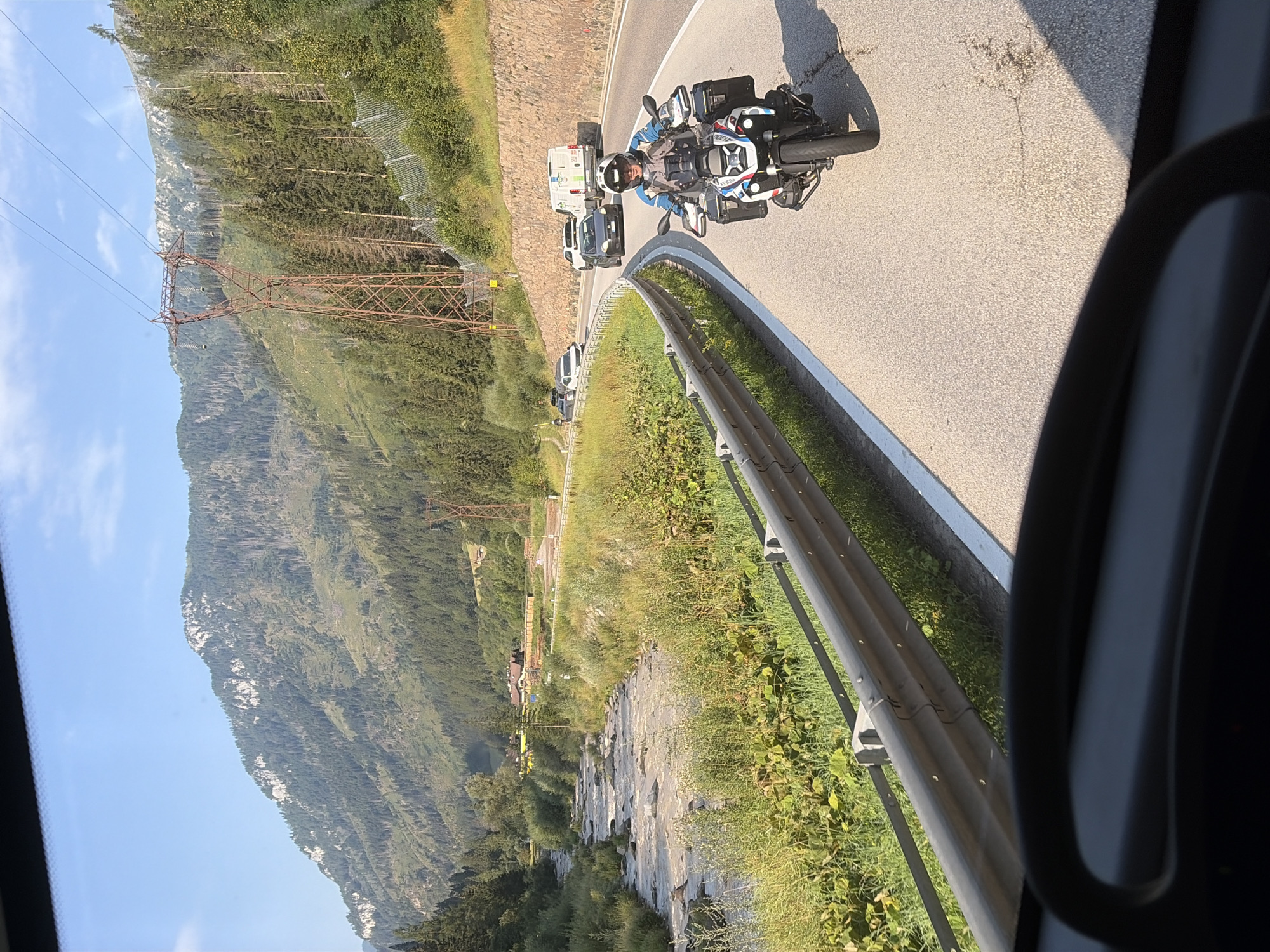 A person is riding a motorcycle on a paved road in a mountainous area. The motorcycle is white, black, and blue, and the rider is wearing a helmet and riding gear. The road curves to the right, and there are several cars and a van on the road ahead. The surrounding environment is a valley with a river, trees, and mountains. The sky is blue with some clouds. The scene appears to be captured from a vehicle moving behind the motorcycle. The location is on the SS48 road leading out of Predazzo, Italy, in the direction of the Rolle Pass.