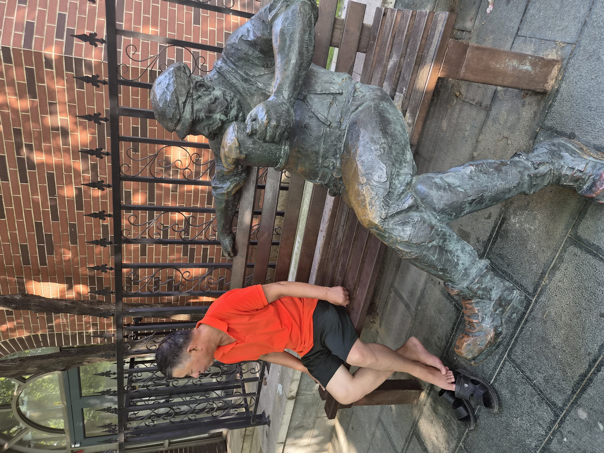 A man in an orange polo shirt and dark shorts sits on a wooden bench next to a bronze statue of a man holding a beer mug. The man is barefoot with sandals on the ground next to him. The statue is weathered and appears to be a public art installation. The background consists of a brick building and a black metal fence. The scene takes place outdoors on a paved area with sunlight casting shadows.