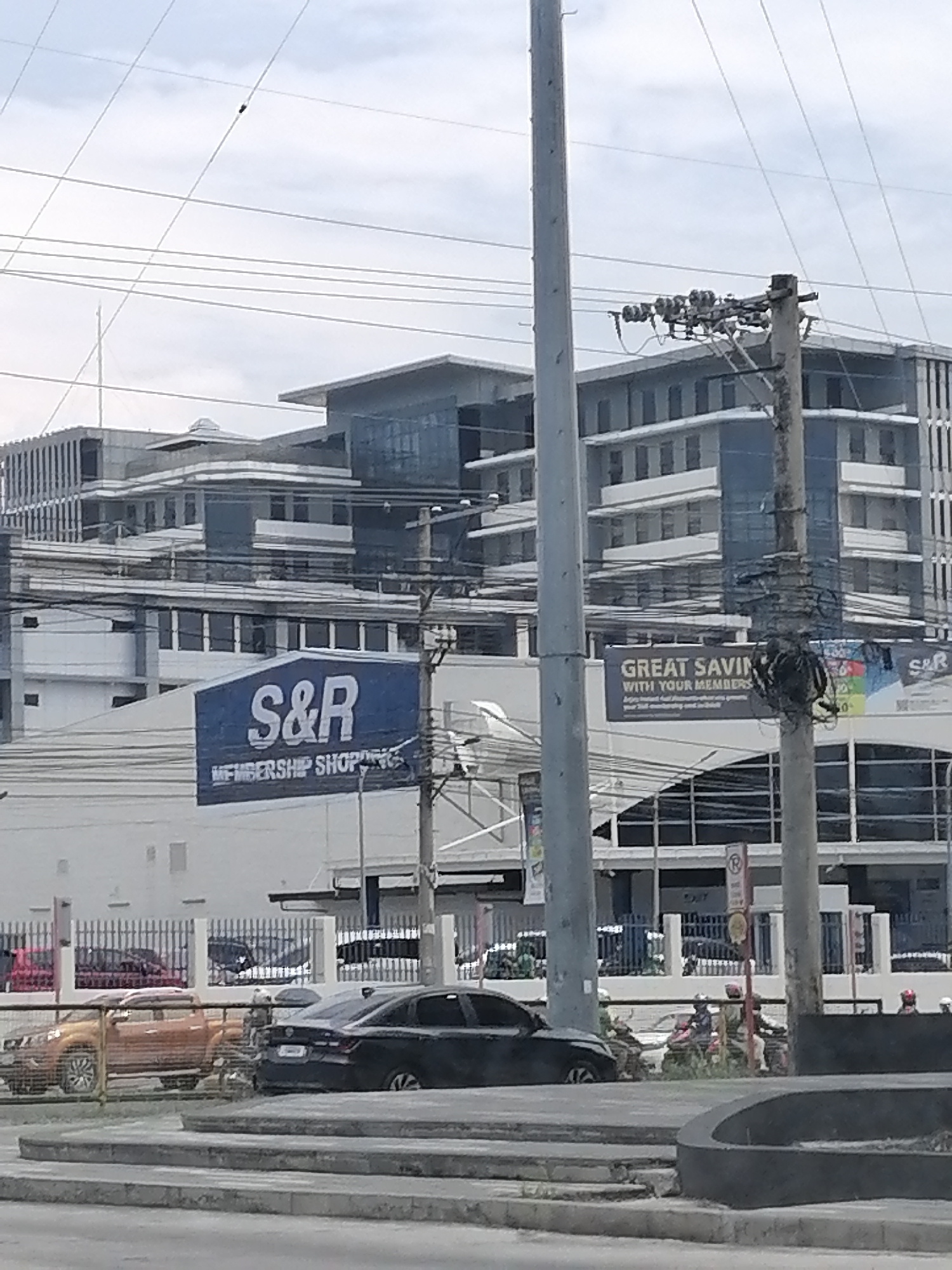 The image presents a daytime urban street scene in Davao City, Philippines, under a diffused, overcast sky. The foreground is dominated by a complex web of overhead power lines strung between several tall utility poles, partially obscuring the view of the buildings behind.
In the midground, a prominent blue and white sign reads "S&R MEMBERSHIP SHOPPING," indicating a large retail warehouse store. To the right of this, another sign with the S&R logo states "GREAT SAVINGS WITH YOUR MEMBERS," with smaller, partially legible text mentioning "Enjoy instant fuel discounts! Save P2/liter on gasoline..."
At street level, several vehicles are visible. An orange pickup truck (resembling a Nissan Navara) is on the left, followed by a black sedan. On the right side of the road, multiple individuals are seen on or near motorcycles. A white fence runs along the roadside, separating it from the commercial establishment. A red circular "NO PARKING" sign is clearly visible, along with the word "EXIT" above a dark barrier.
The background features several multi-story buildings with modern architecture, characterized by glass facades and light-colored exteriors. Some sections appear to be newly completed or still under construction, suggesting ongoing urban development.
The scene depicts a commercial hub with active traffic and typical Southeast Asian urban infrastructure, including the dense network of power lines. The presence of the S&R store and the vehicle types are consistent with a bustling area in the Philippines.