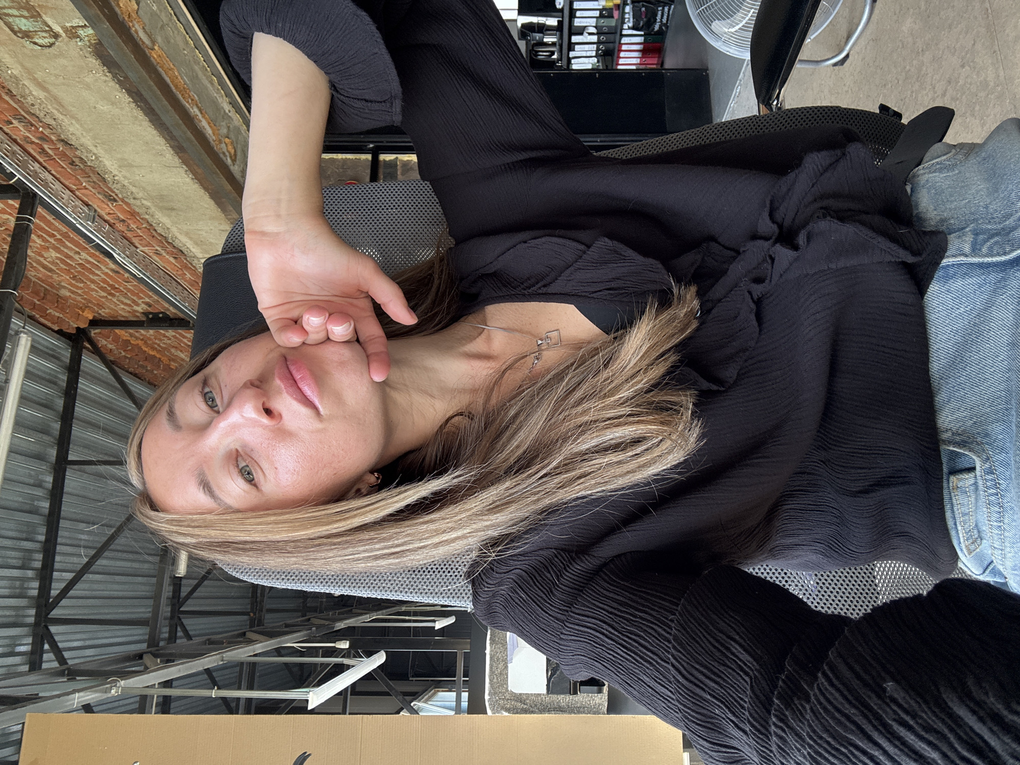 A woman with blonde hair is seated in an office chair, looking towards the camera. Her right hand is resting on her chin. She is wearing a black blouse with ruffled details and blue jeans. The background suggests an industrial-style office with exposed metal beams on the ceiling and brick walls. The location in the city of Chaikovsky, Russia is not discernable from the visual information provided.