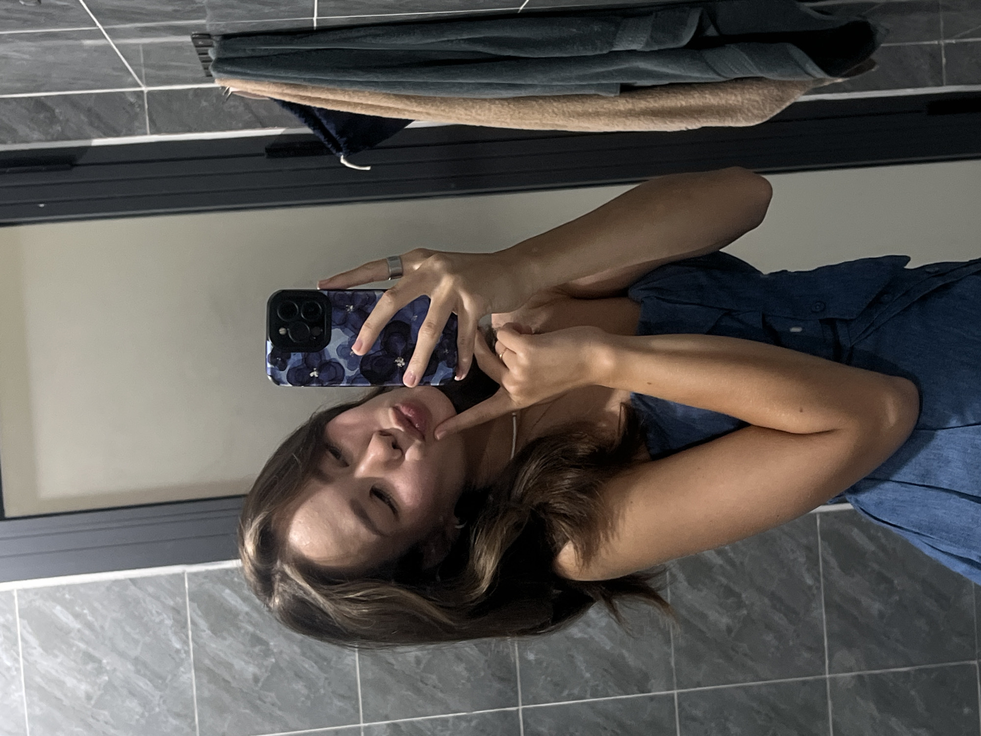 A woman is taking a selfie in a bathroom with grey tiled walls and floor. She is holding a phone with a blue flowered case. The woman is wearing a denim shirt. There are towels hanging on a rack above a mirror. The location appears to be a bathroom, possibly in Nam Nha Trang Ward, Vietnam.