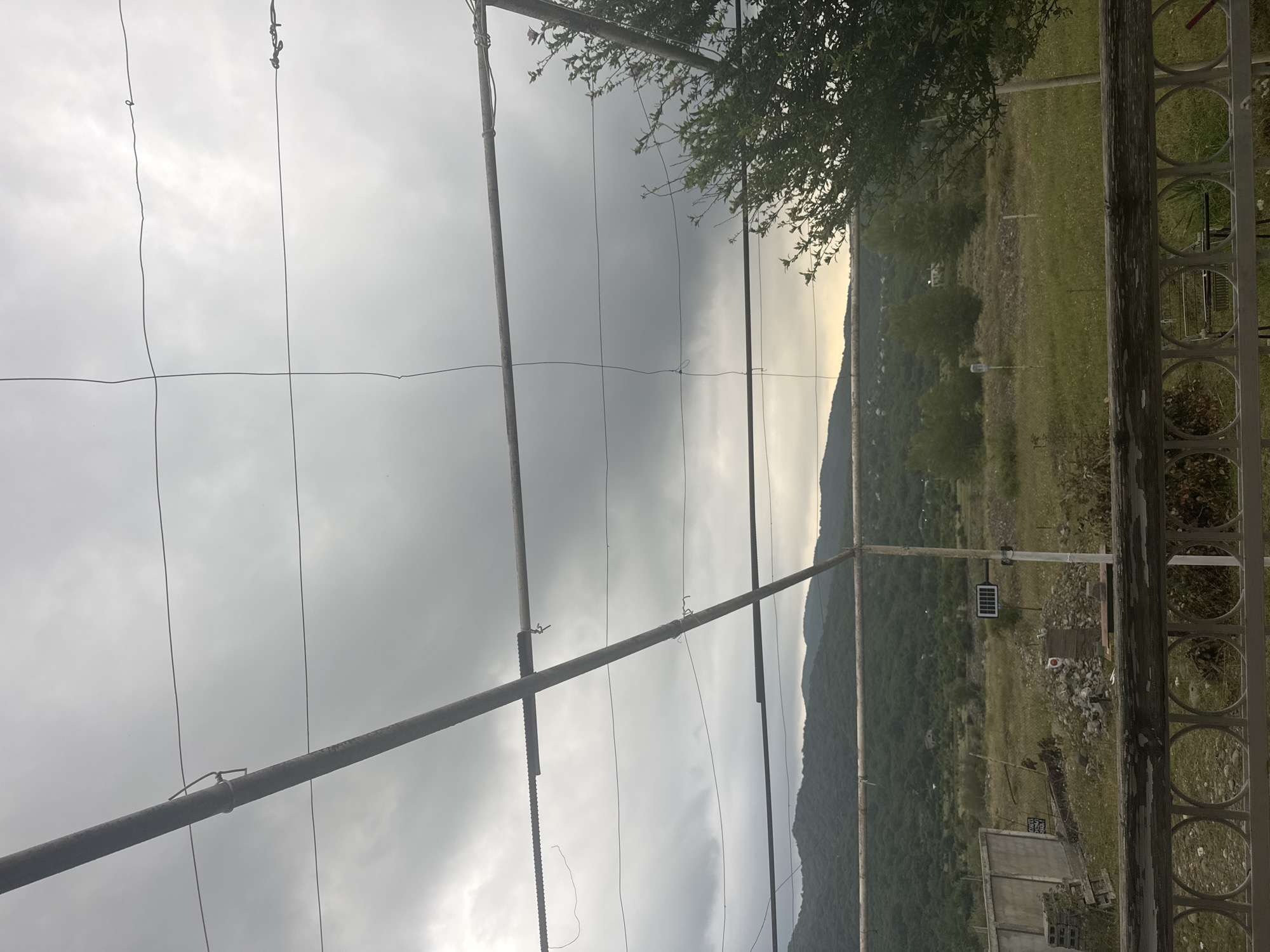 The image depicts an outdoor scene in Aragvispiri, Georgia, under a cloudy sky. In the foreground, a weathered wooden railing with circular metal accents separates the viewer from the rest of the scene. Above the railing, a metallic structure composed of horizontal and diagonal beams supports thin wires that crisscross the frame.
The middle ground features a landscape with a gently sloping hill covered in lush green trees and vegetation. A concrete wall, possibly part of a building or boundary, is visible on the left. Scattered debris, including what appears to be a pallet and rocks, lies on the ground. A small solar-powered light is mounted on a pole in the middle of a field.
The background shows a range of hills and mountains, also densely vegetated, fading into the overcast sky. The overall impression is one of a rural or semi-rural setting. No people are present in the image.