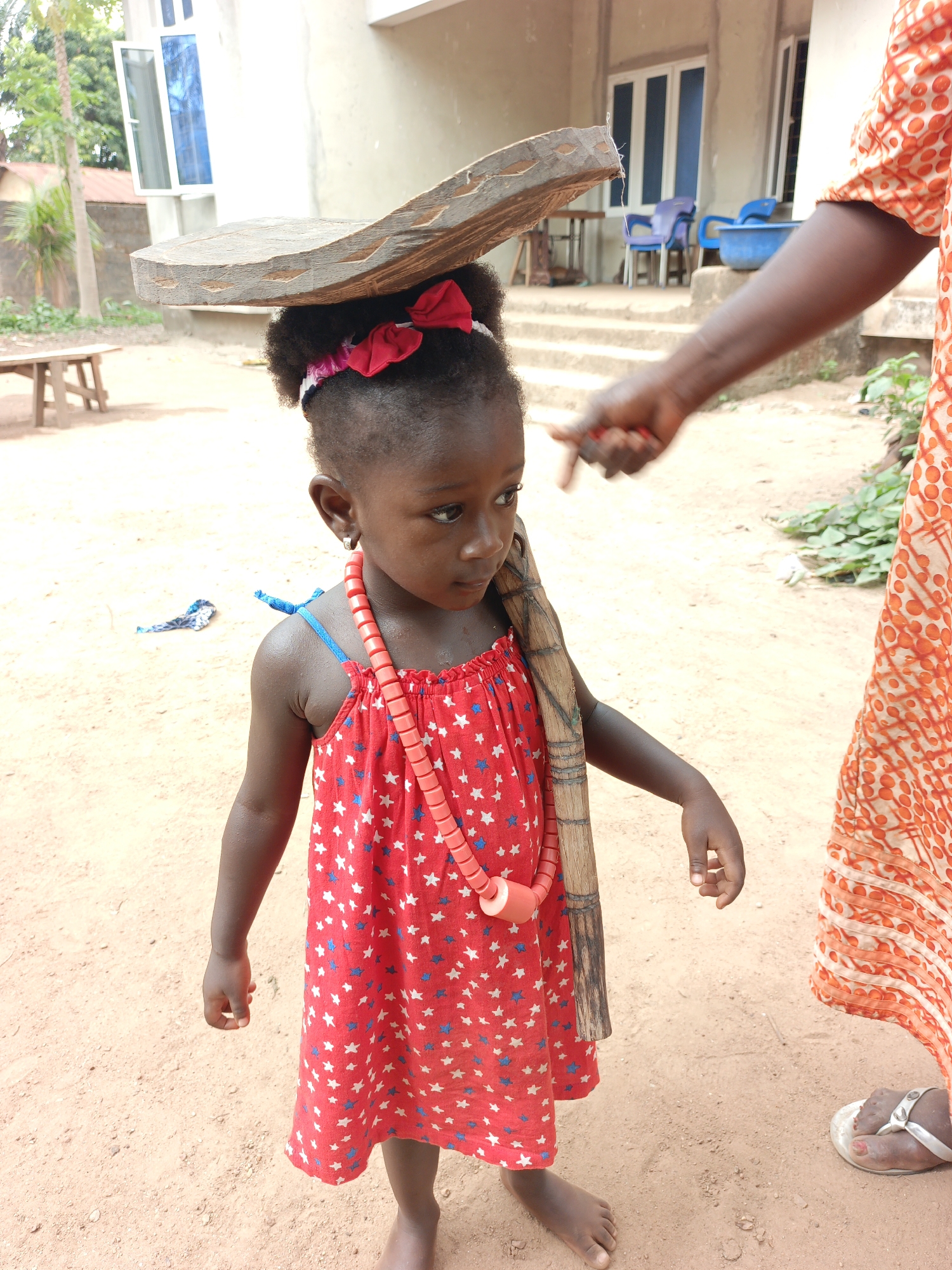 A young, dark-skinned girl stands barefoot in a dirt yard in Akwanga, Nigeria. She wears a red sundress patterned with white and blue stars, a pink and white headband with red bows, and a coral-colored necklace. She is balancing a flat, roughly hewn wooden object on her head and holding a long, thin, light-brown wooden object on her shoulder. An adult's arm, clad in an orange patterned dress, extends into the frame from the right. The adult is wearing a silver sandal. In the background is a single-story white building with blue-trimmed windows. A small wooden bench and blue plastic chairs are also visible.