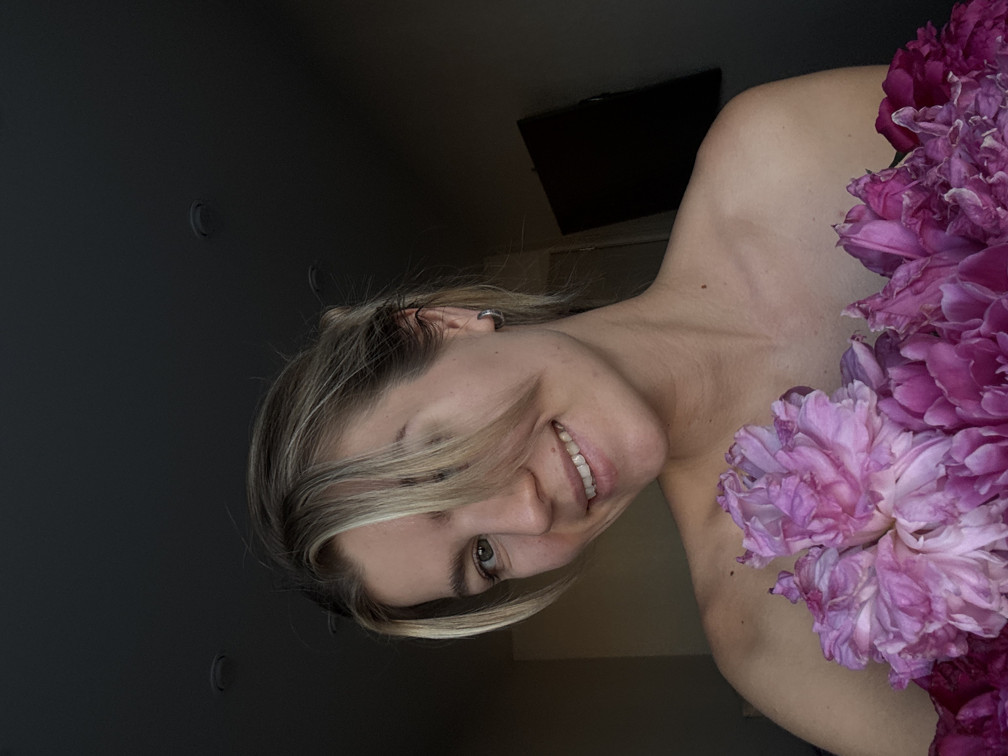 A young woman with blonde hair, wearing a silver hoop earring, is pictured in a close-up, bust-up shot, holding a bouquet of pink peonies. She is looking towards the viewer with a slight smile. The background is dark with a black television screen visible on the right wall and two circular light fixtures on the ceiling. The woman's shoulders and upper chest are bare.