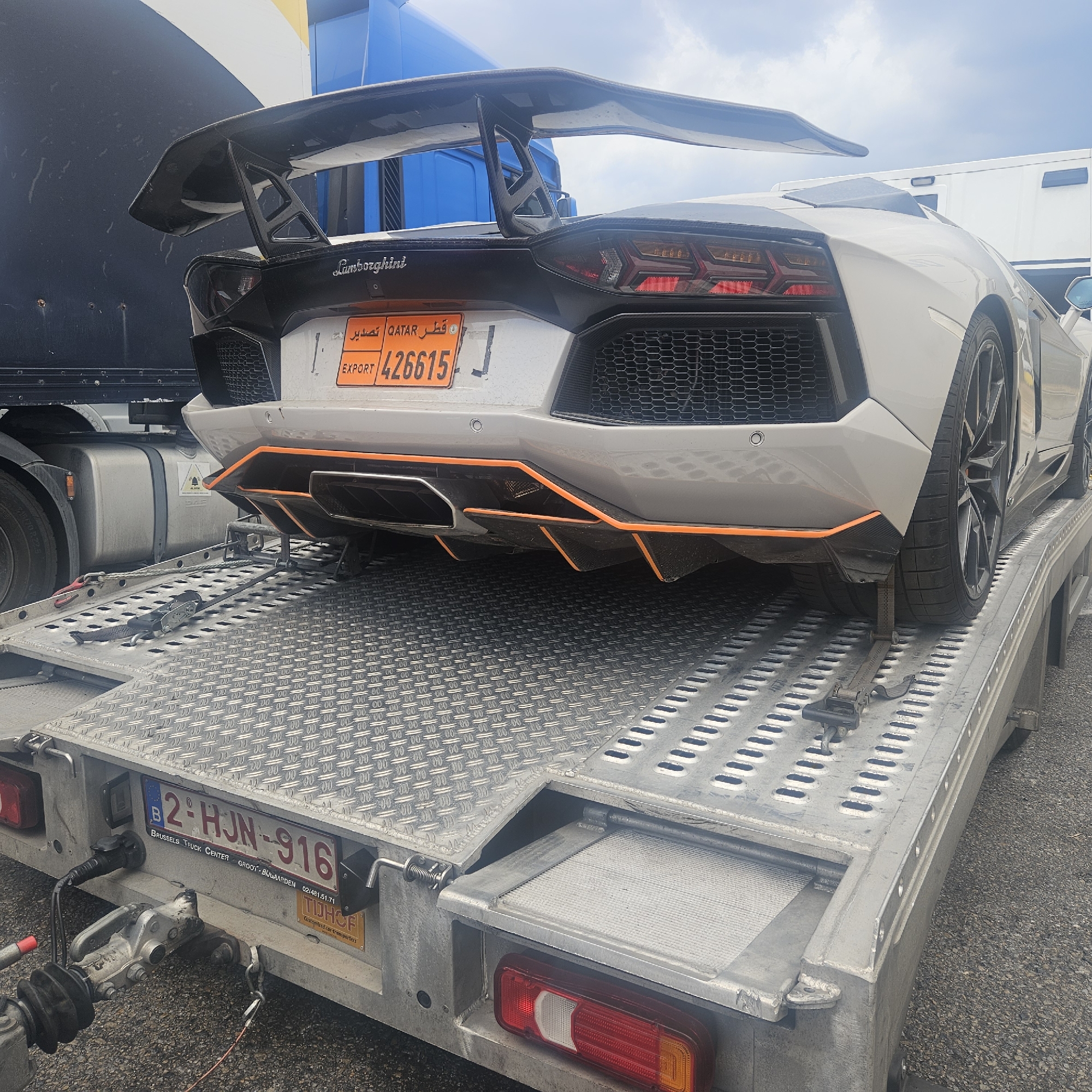 A white Lamborghini Aventador is being transported on a flatbed trailer. The car has a large rear spoiler and an orange accent on its diffuser. The license plate on the car is orange with "EXPORT" written on it and the number "426615". The trailer has a Belgian license plate that reads "2°HJN-916". The trailer is attached to a truck, and the scene appears to be outdoors on a paved surface. The location is identified as La Jonquera, Spain, a border town known for its commercial activity. The sky is cloudy, and the lighting suggests it is daytime. There are no people visible in the image. The trailer is equipped with straps to secure the vehicle during transport.