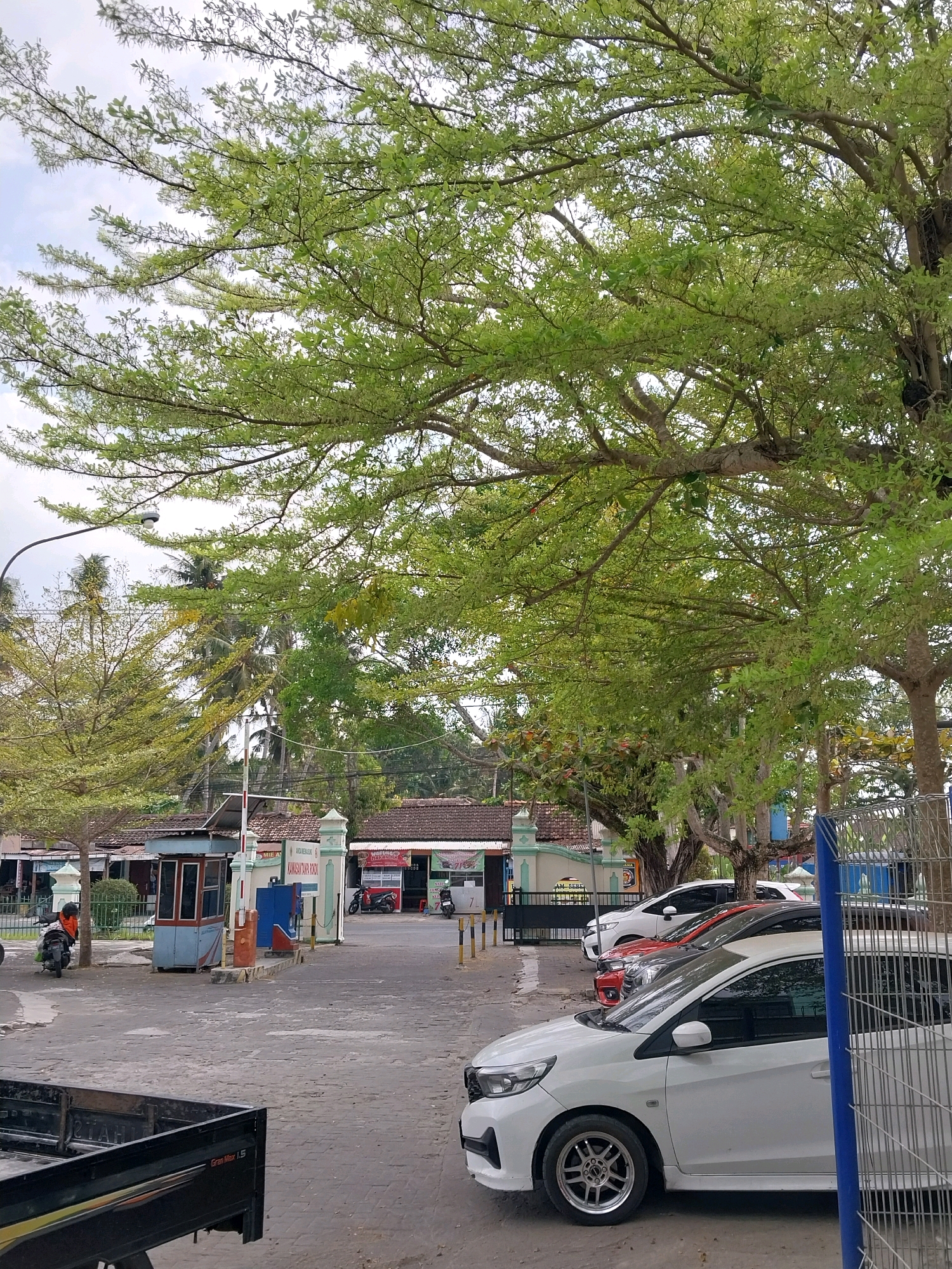 In the city of Wates, Indonesia, a gray, paved road is visible, bordered by a black pick-up truck on the left and parked white cars on the right. In the background, buildings with red tile roofs line the street, and a guardhouse stands near the entrance. Lush green trees with sparse leaves dominate the upper portion of the image, their branches extending over the scene. The sky above is overcast. Two people are seen on motorbikes, one in an orange jacket, and several other motorbikes are parked near the buildings. A blue fence is partially visible on the far right. The overall impression is that of a sunny day in a tropical urban environment.