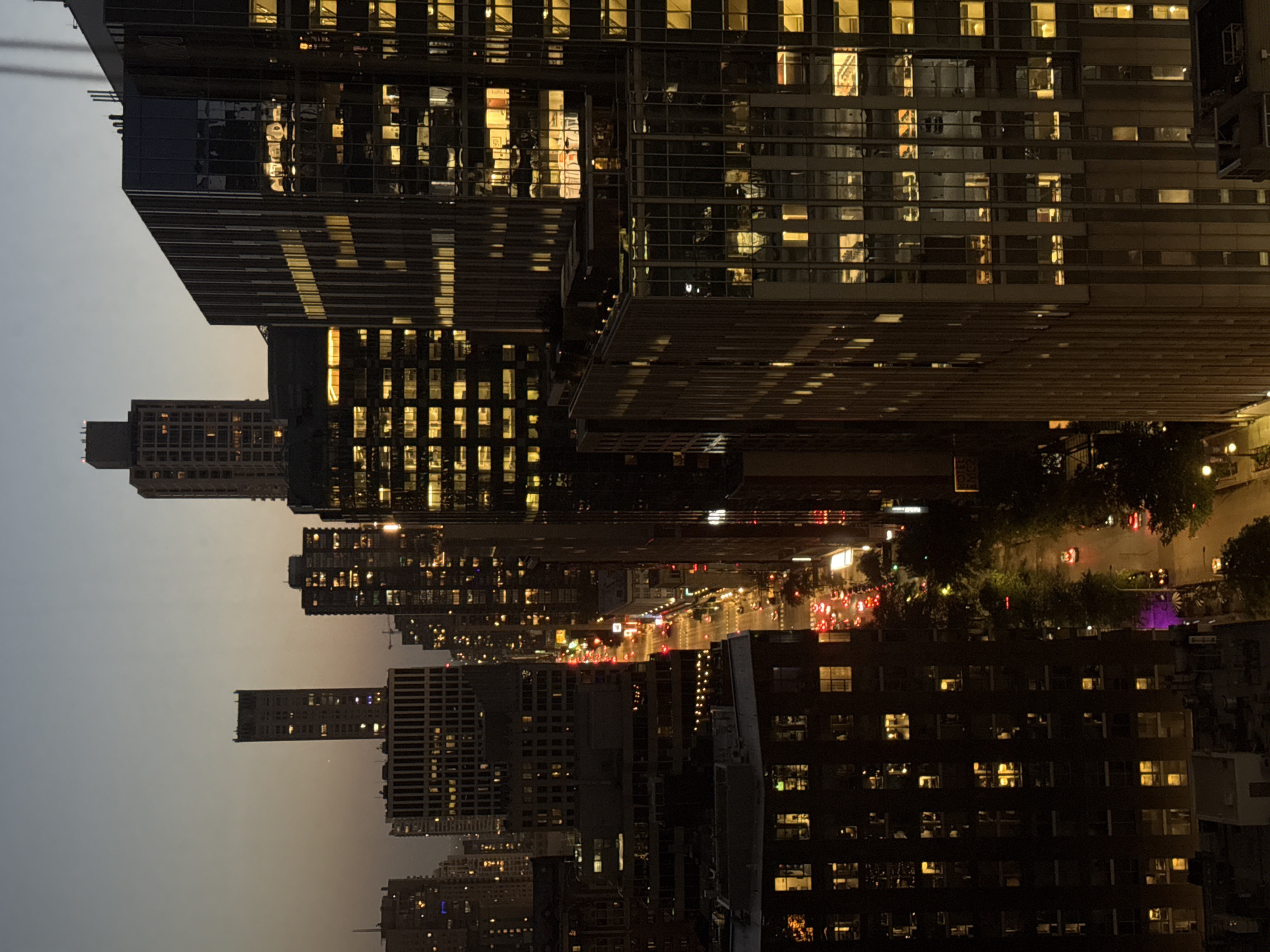 An elevated, long shot captures a dusk aerial view of a city street in Chicago, USA. Tall, illuminated skyscrapers line both sides of a street with a moderate flow of traffic. The buildings are a mix of modern glass structures and older brick buildings, all aglow with interior lights. The sky is a dusky gradient of grey and orange. The street below is visible, with cars and streetlights casting a warm glow on the wet asphalt, suggesting recent rain. Trees line the sidewalk, their leaves dark silhouettes against the illuminated buildings. In the foreground, the tops of lower buildings are visible, their windows also lit. The overall impression is one of urban vibrancy and the transition from day to night.