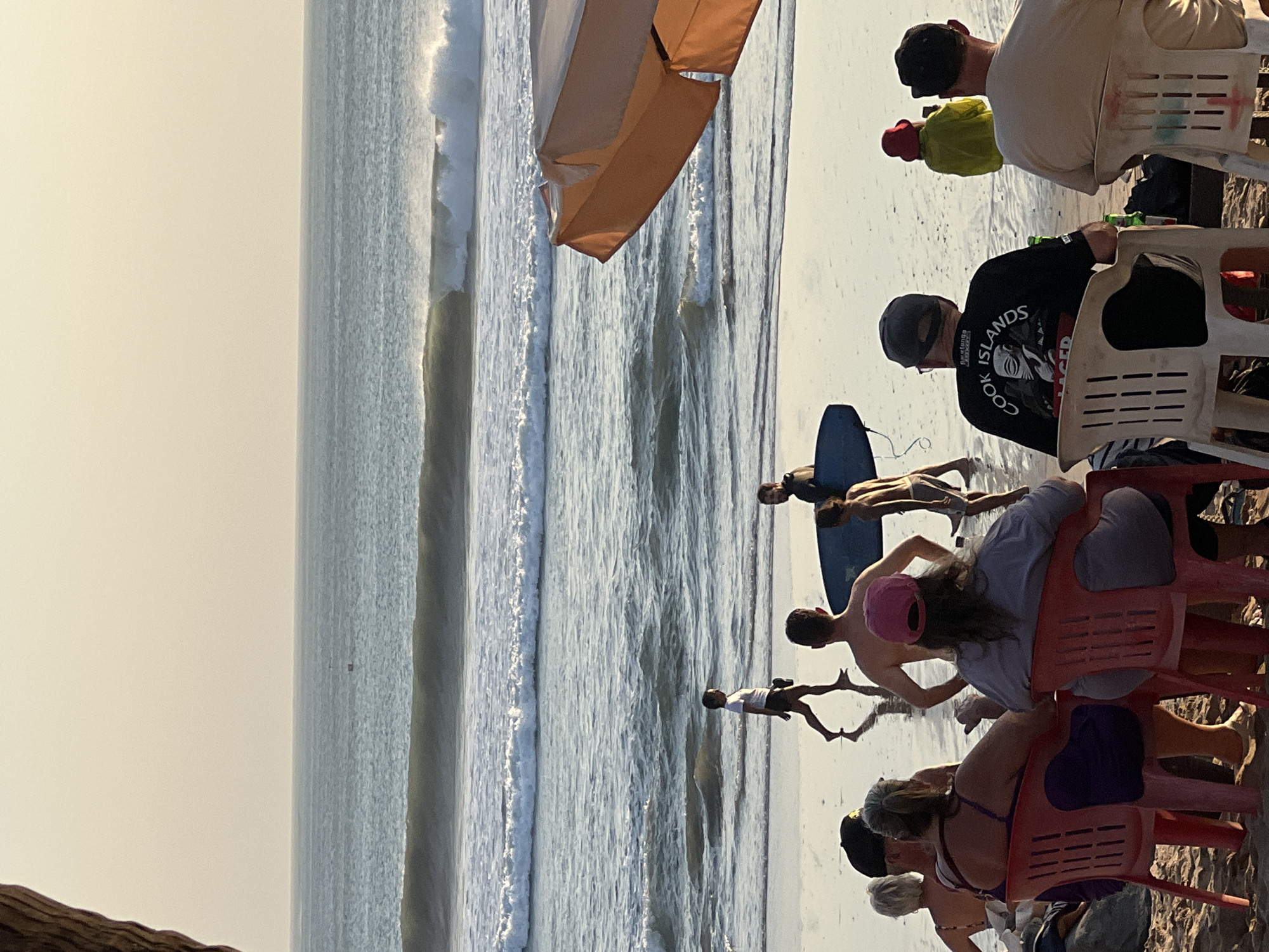 An overhead view captures a serene beach scene in Kuta, Indonesia, during the golden hour. The sky, a soft gradient of pale yellow and pink, meets the tranquil ocean. Gentle waves roll onto the shore, leaving wet sand that reflects the soft light. In the foreground, a group of people are seated on chairs facing the ocean, some appearing to be tourists, others locals. Two people are closer to the water, one standing and the other carrying a surfboard. The overall atmosphere is peaceful and relaxed, a typical Kuta beach experience.