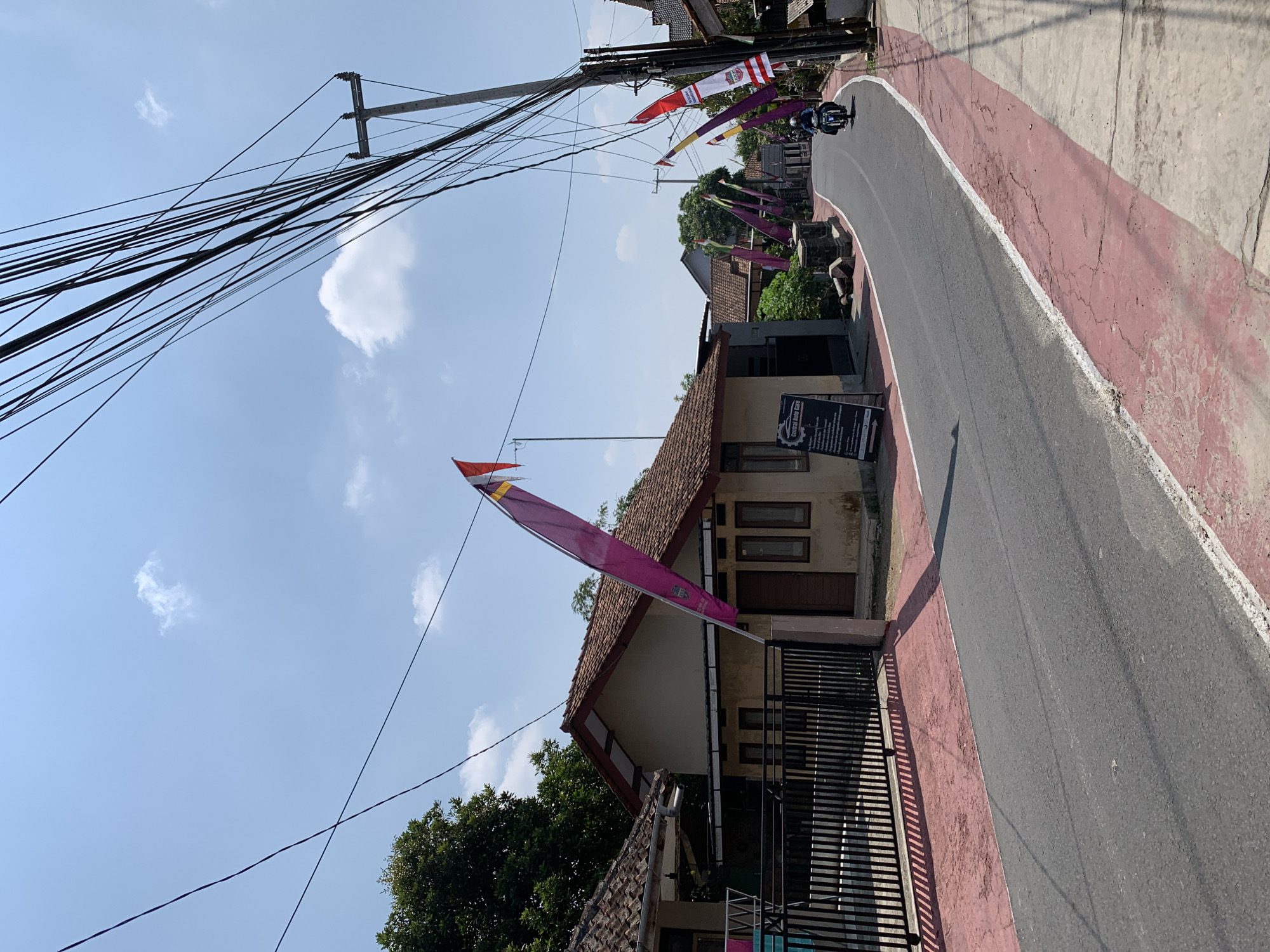 A street scene in Ciamis, Indonesia, shows a road lined with houses and flags under a clear sky with scattered clouds. A motorcycle is visible on the road, moving away from the viewer. The road has a red border. The houses have tiled roofs and one has a dark metal fence. Electrical wires are strung overhead.