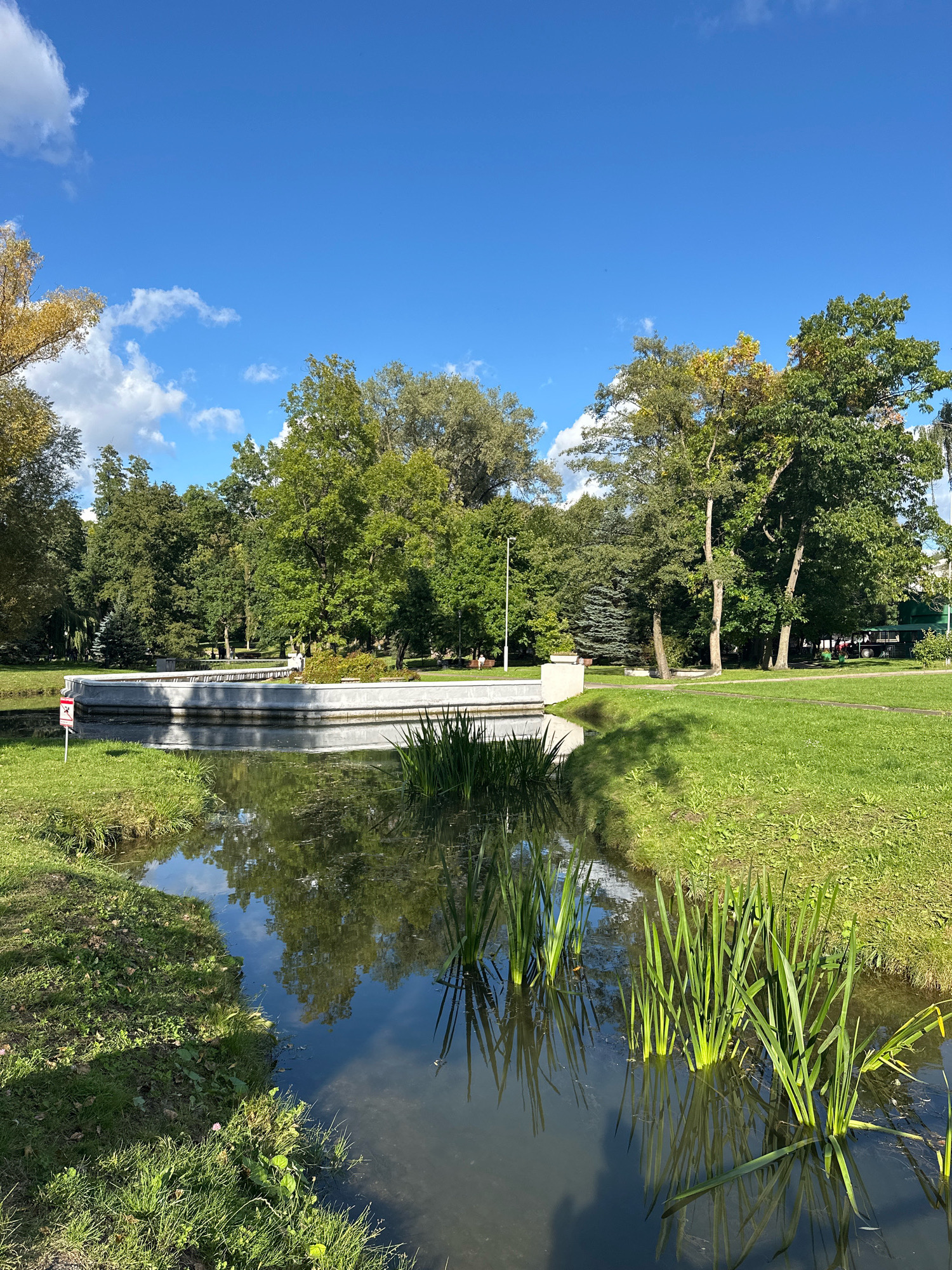 A serene view of a park in Minsk, Belarus, on a clear day, features a calm body of water in the foreground, reflecting the blue sky and surrounding greenery. Tall, lush green plants emerge from the water, their reflections creating a symmetrical pattern. The water's edge is lined with manicured green grass. In the middle ground, a circular water feature with a low, light-colored concrete rim is visible, with more green grass and trees in the background. The trees exhibit a mix of green and autumnal yellow foliage, suggesting the season. A few people are scattered throughout the park, engaged in leisurely activities. The overall atmosphere is tranquil and picturesque.